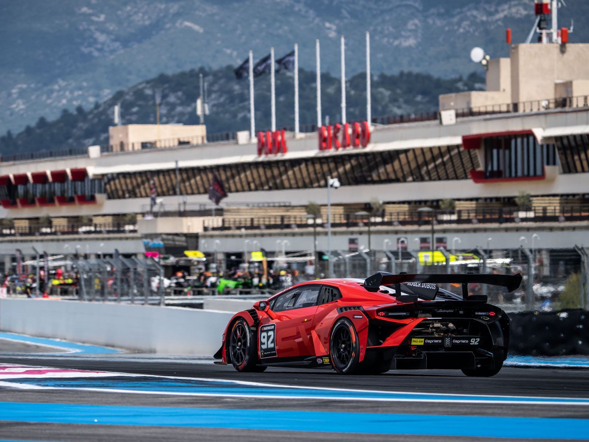 A red lamborghini huracán super trofeo evo2 race car seen from the rear three-quarter angle on track, showing the large carbon-fiber rear wing and diffuser