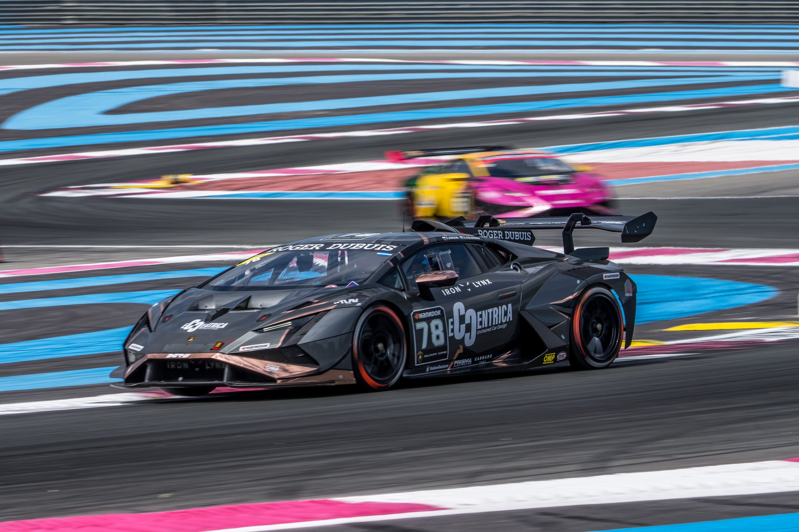 A dark grey lamborghini huracán super trofeo evo2 speeds around a corner with blurred competitors visible in the background