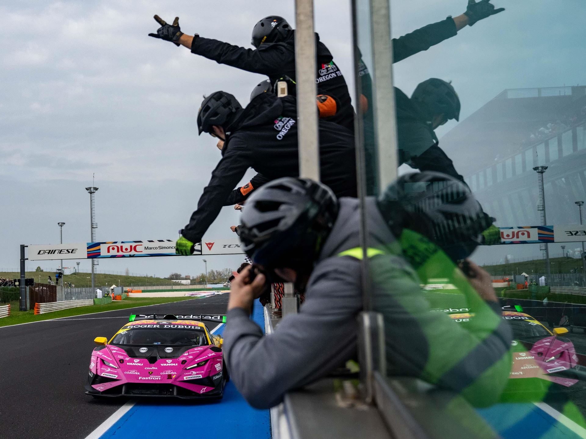A pink and yellow lamborghini huracán super trofeo evo2 passes the pit wall as crew members cheer