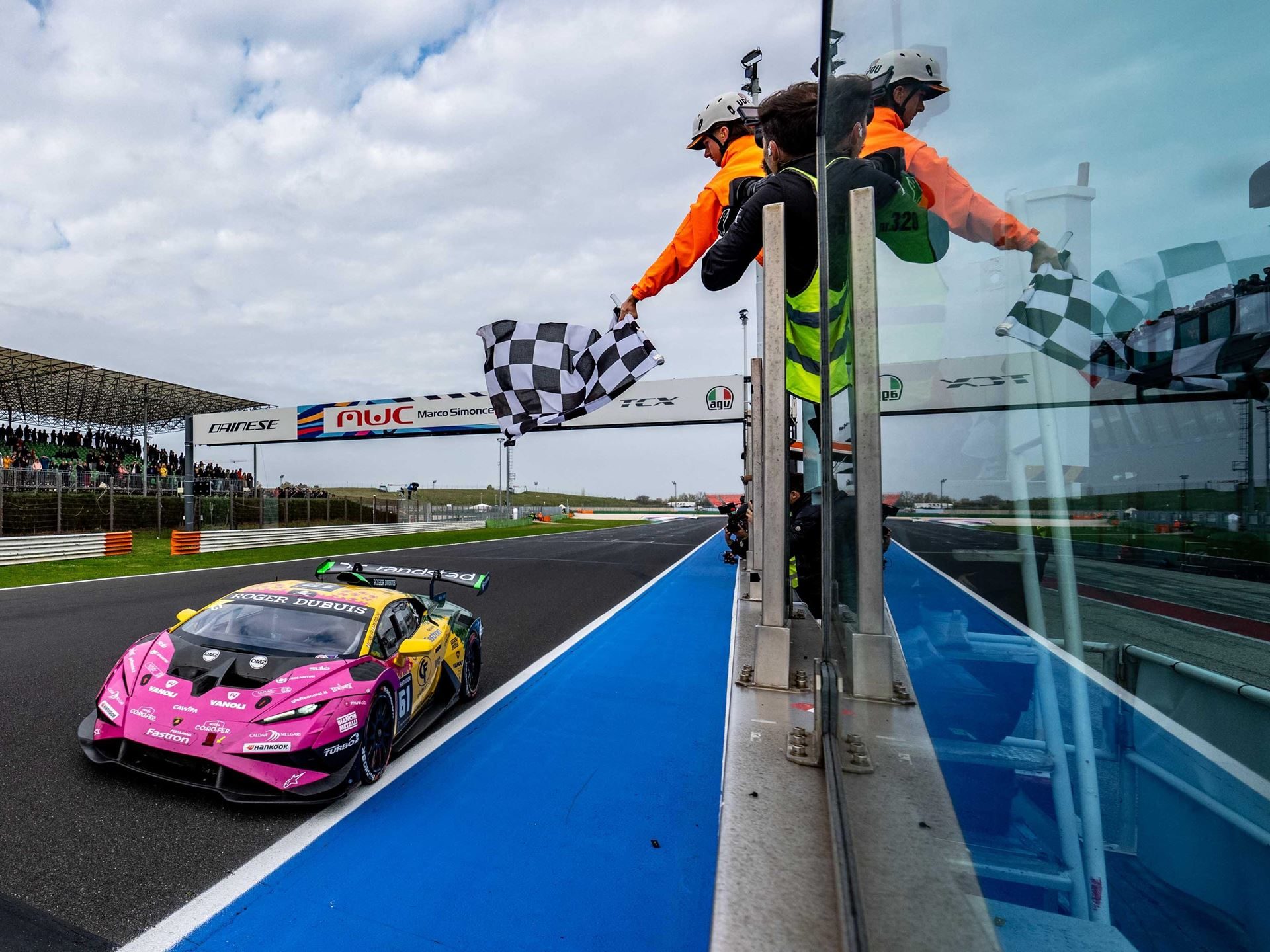 A pink and yellow lamborghini huracán super trofeo evo2 crosses the finish line under the checkered flag at misano