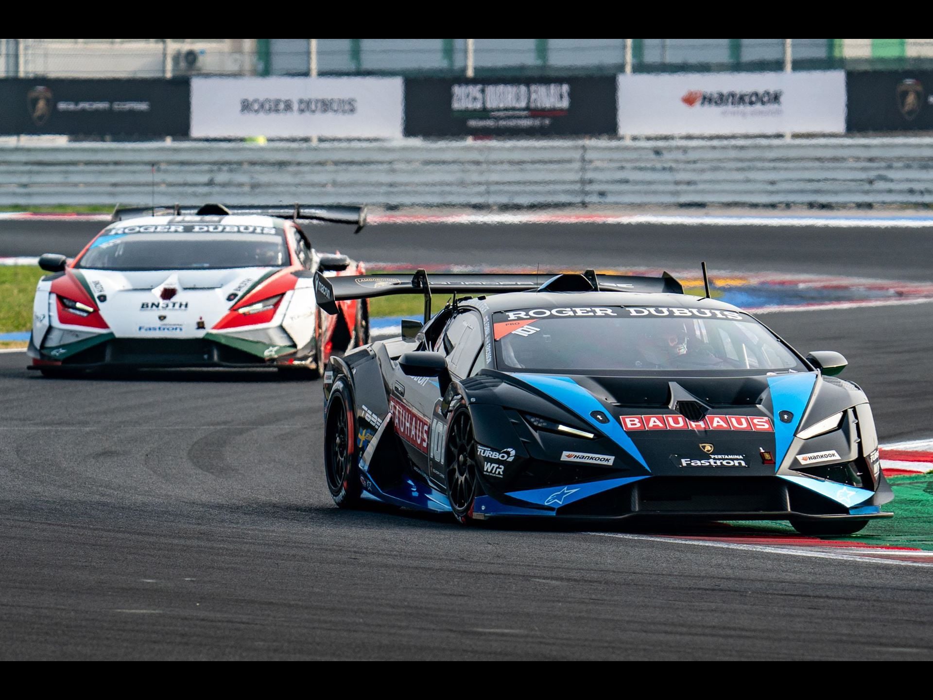 One-Point Margins And Last-Lap Passes: How The 2025 Super Trofeo Season Built The Most Dramatic World Finals In Years 7 Black and blue lamborghini huracán super trofeo evo2 leads a white and red car on track with 2025 world finals banner visible
