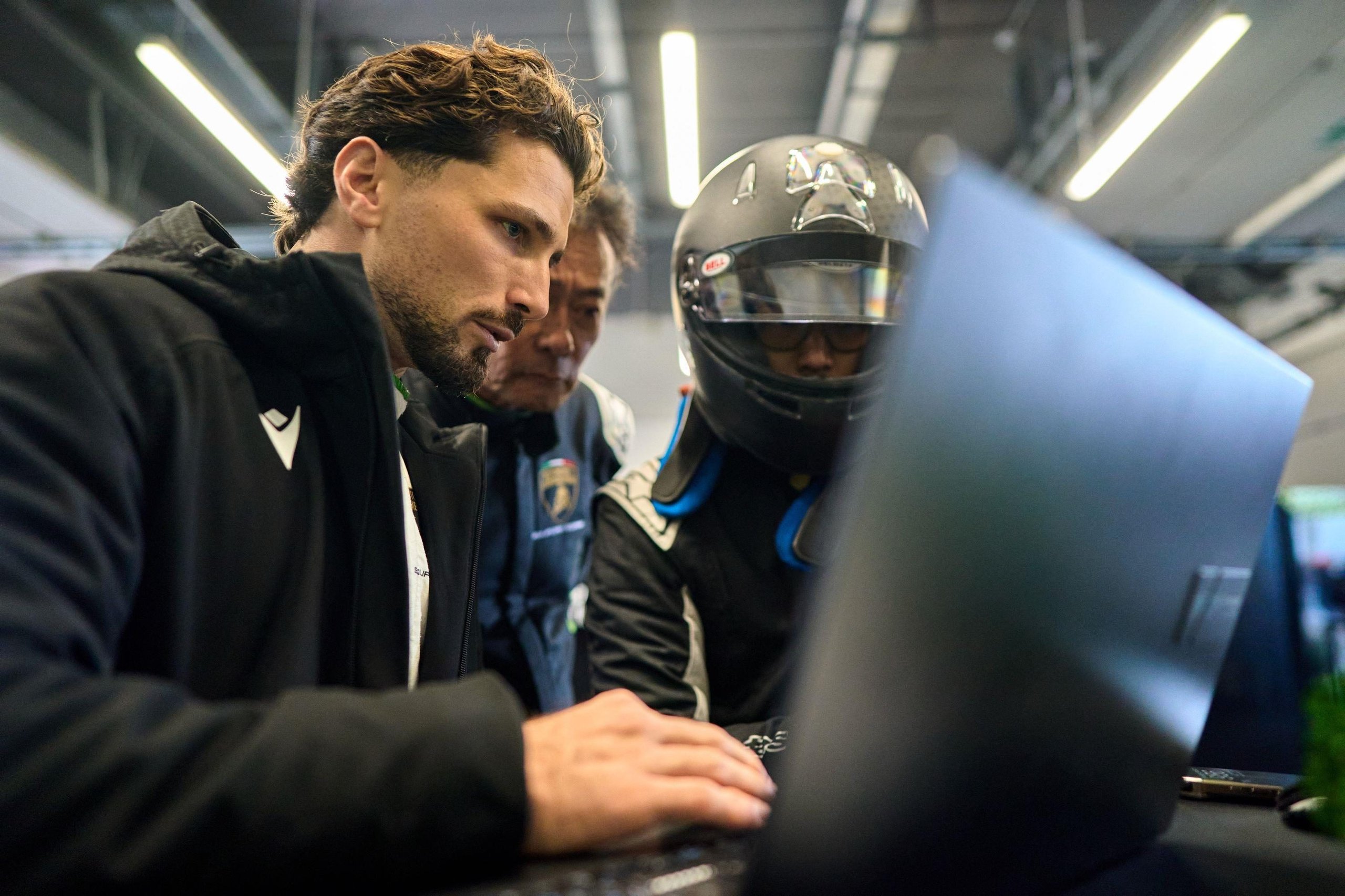 Four Weekends, 40 Cars, Factory Drivers: Inside Lamborghini'S 2026 Essenza Scv12 Program 7 Three individuals including a helmeted driver gathered around a laptop in a garage setting reviewing telemetry data