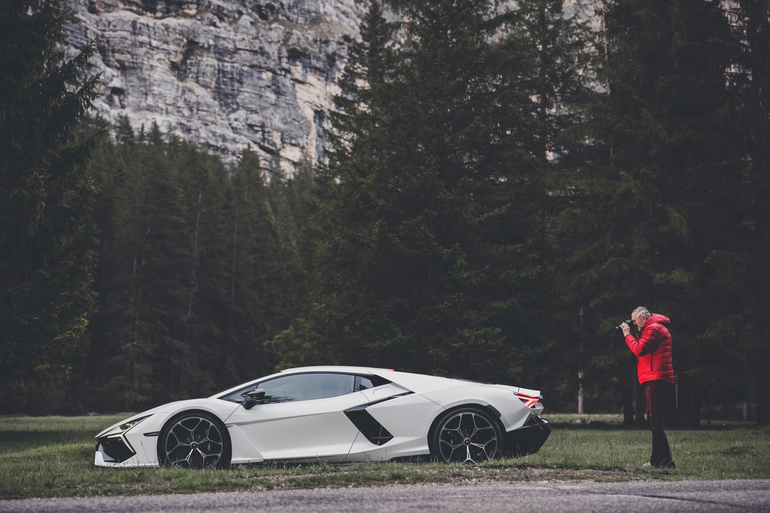 Anton Corbijn Photographs The Revuelto Like A Rock Star, And That'S Exactly Lamborghini'S Point 3 Anton corbijn in a red jacket photographing a white lamborghini revuelto against a forest and rocky cliff backdrop in the dolomites