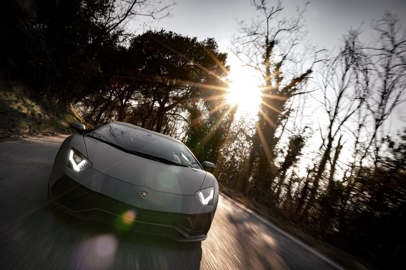 Grey Lamborghini Aventador Ultimae driving on a winding road at sunset with lens flare through trees