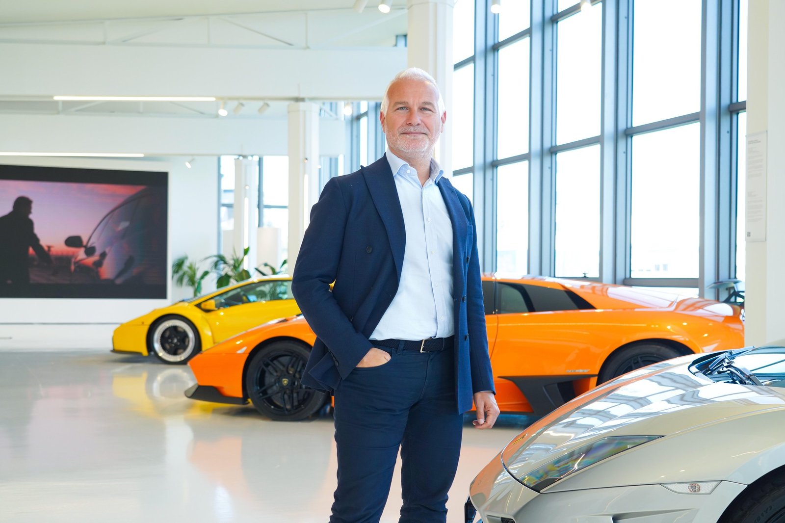 Christian Mastro standing in a Lamborghini showroom with yellow, orange, and silver models behind him