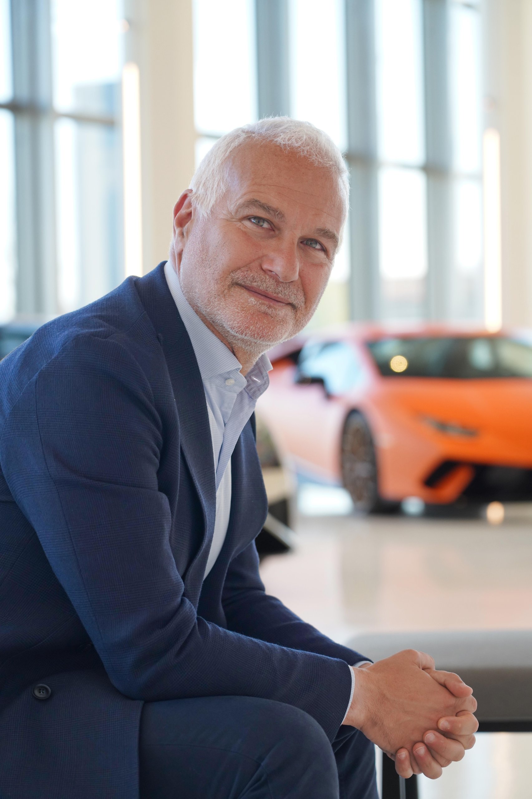 Christian mastro seated in a showroom with an orange lamborghini huracán in the background