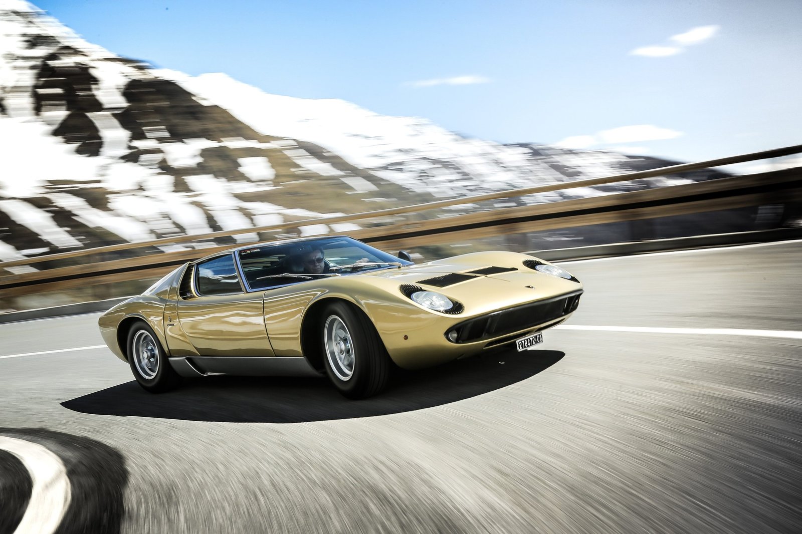 A gold Lamborghini Miura P400 driving dynamically on a winding mountain road with snow-capped peaks in the background