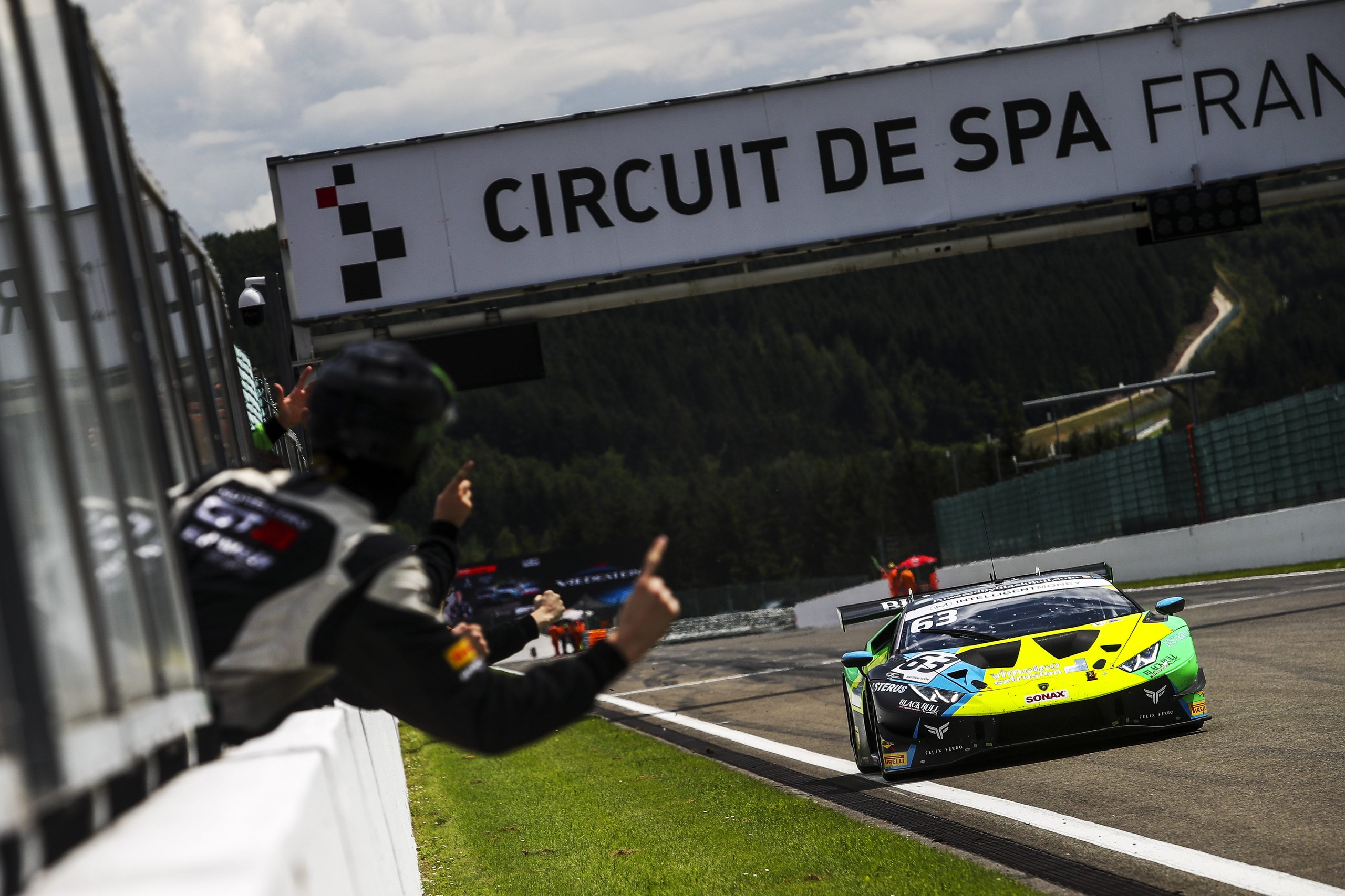 The #63 lamborghini huracán gt3 evo in green and blue livery racing past a trackside official at spa-francorchamps