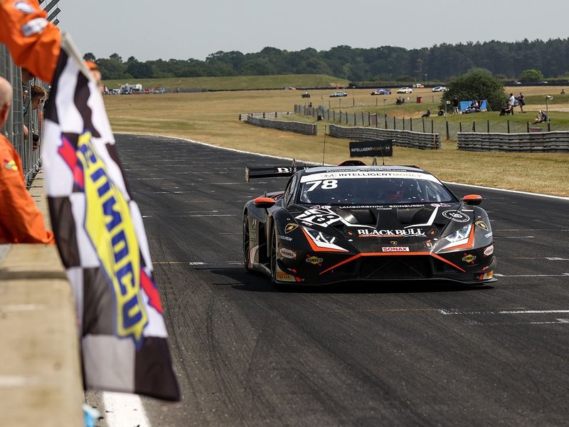 Black Lamborghini Huracán GT3 EVO2 with orange accents crossing the finish line under a checkered flag at a sunny racetrack