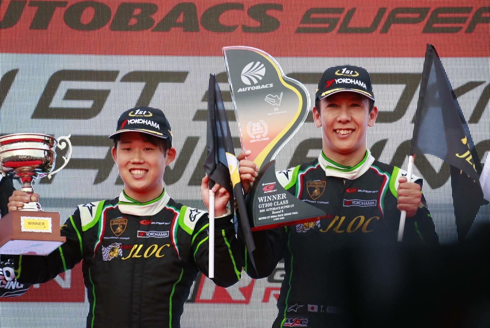 Jloc drivers takashi kogure and yuya motojima on the podium holding trophies after winning the 2023 super gt season finale at motegi
