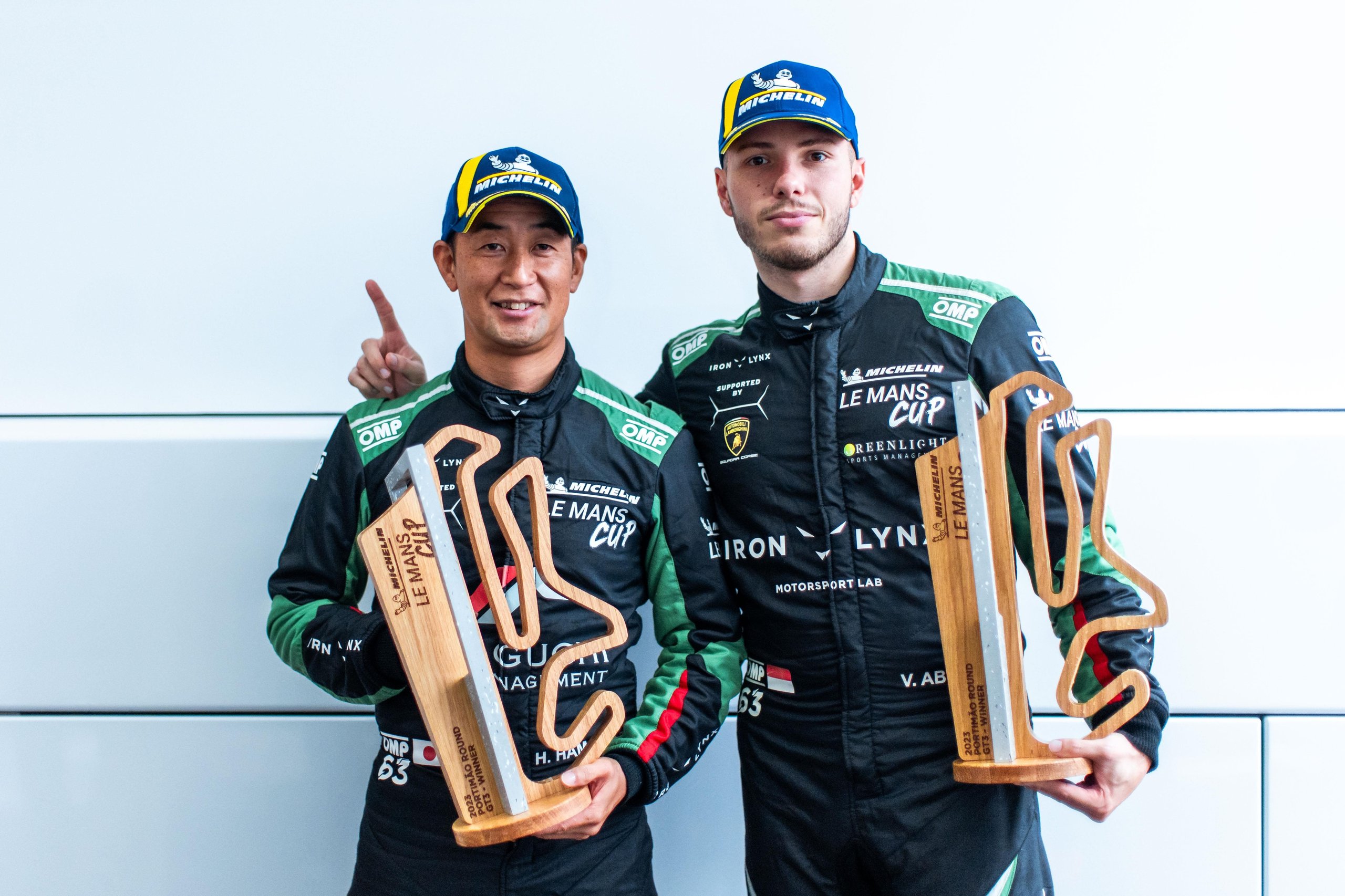 Hiroshi hamaguchi and vincent abril in black racing suits and michelin caps celebrating with le mans cup trophies after iron lynx's first victory at portimão