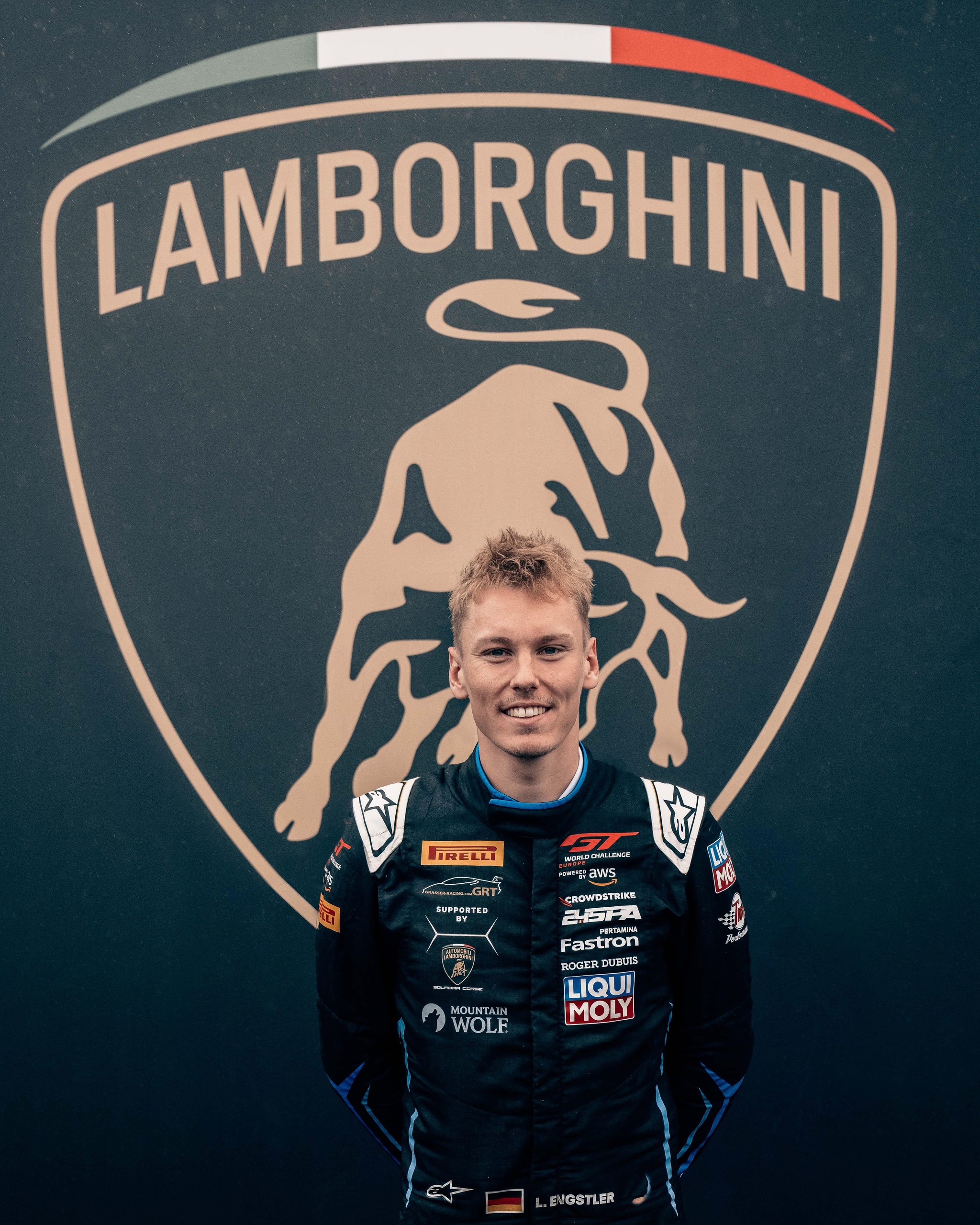 Young Lamborghini factory driver in black racing suit smiling in front of a large Lamborghini logo backdrop