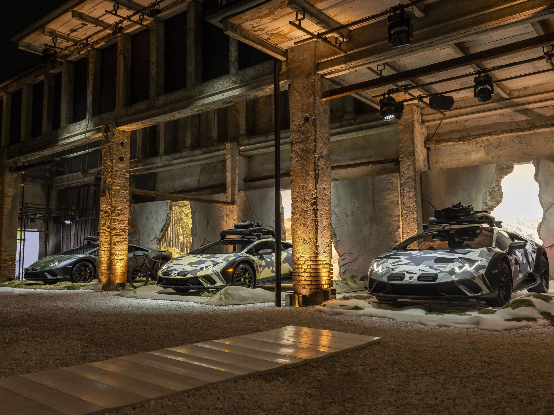 Twelve Camouflaged Sterrattos, A Michelin-Starred Dinner, And Tod'S Loafers: Inside Lamborghini'S Milan Design Week Power Play 3 Three huracán sterrato all terrain models displayed in an industrial setting with themed desert, forest, and snow backdrops