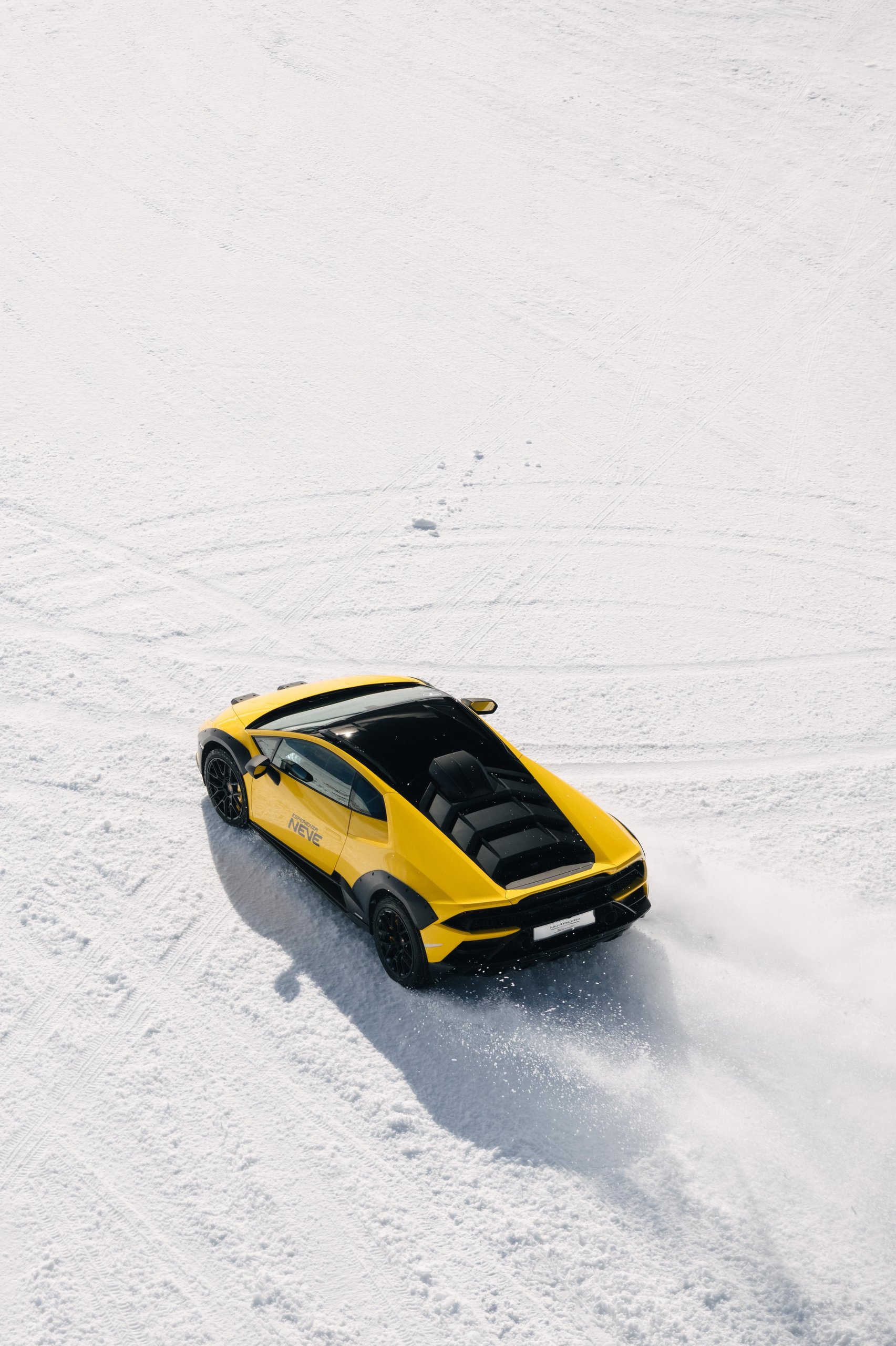 Yellow huracán sterrato driving away on snow with tire tracks in foreground and snow spray from rear