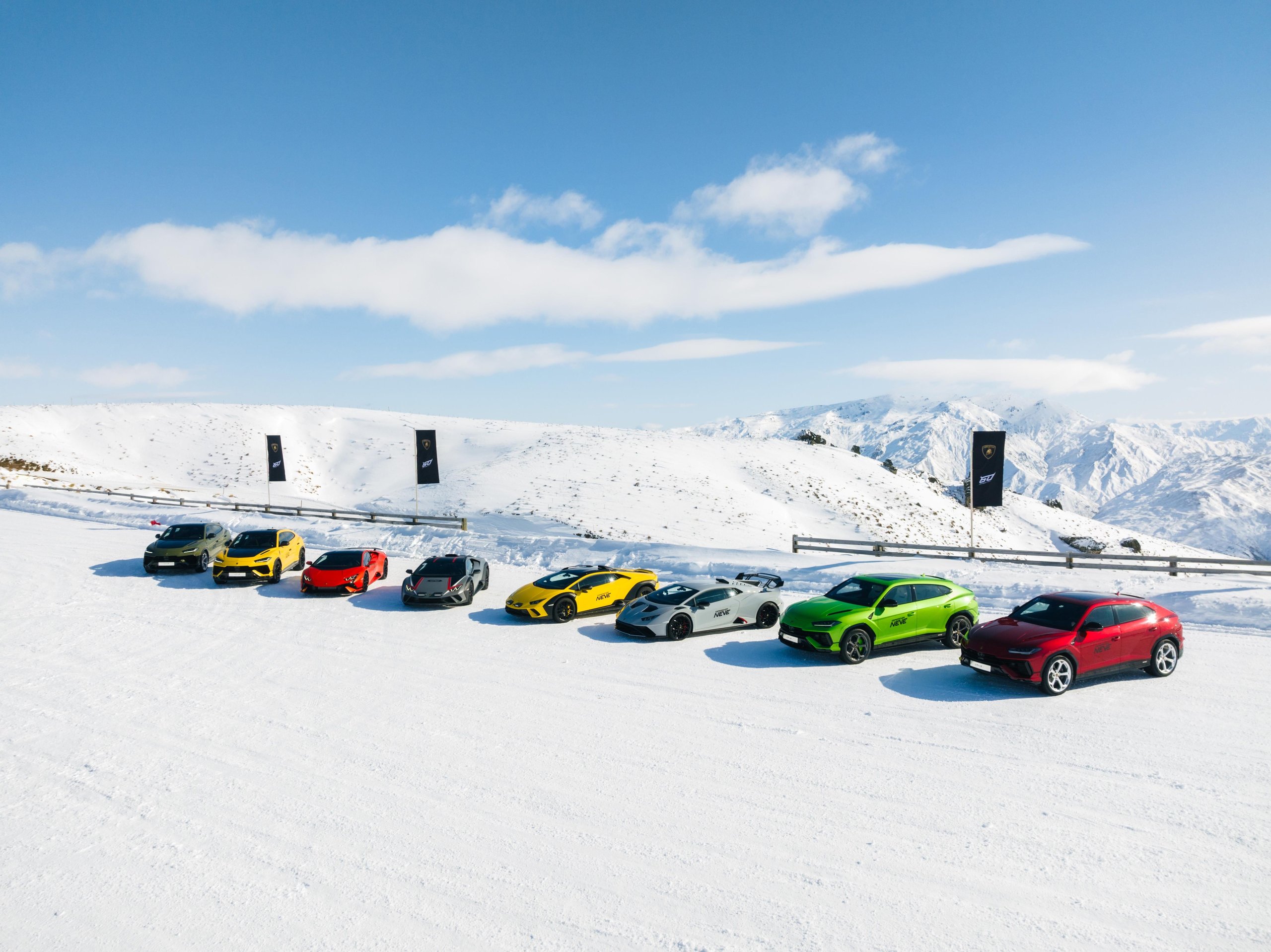 Colorful lineup of lamborghini models on a snowfield with snow-capped mountain range under blue sky