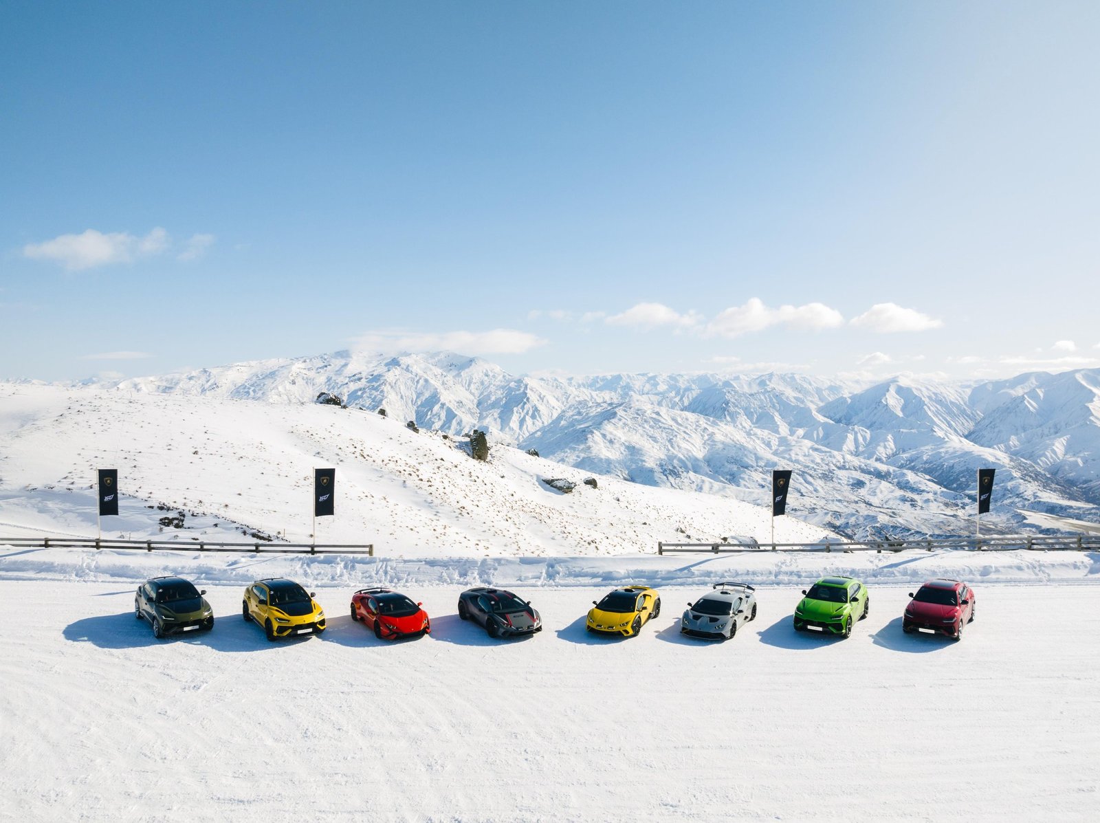 Multiple Lamborghini models lined up on a snow-covered plain in Queenstown, New Zealand, with a mountain range under clear blue sky