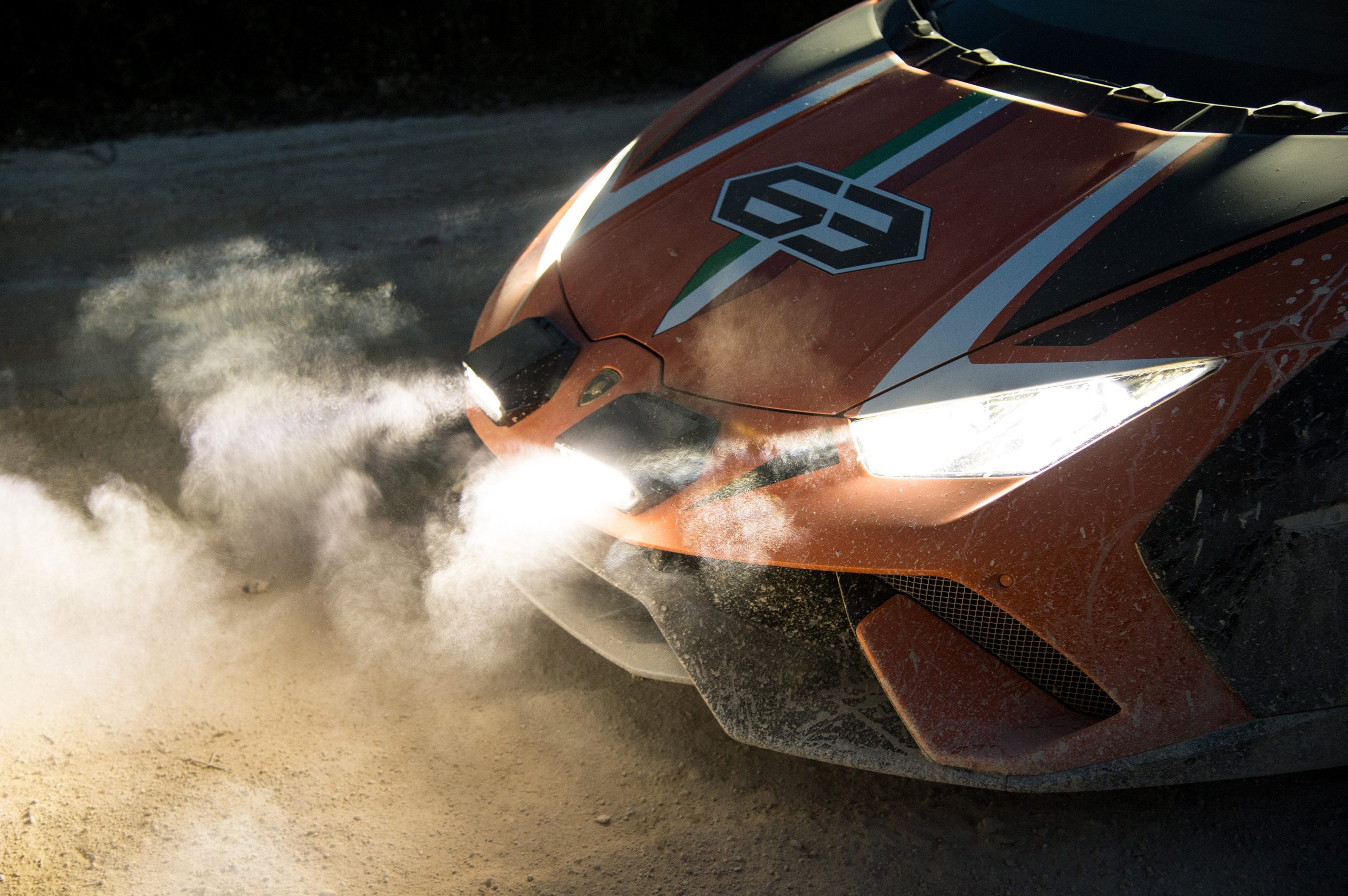 How A Dusty Dinner Conversation And Chassis #53 Gave Birth To The Huracán Sterrato 5 Close-up of the orange huracán sterrato concept's front end with illuminated auxiliary lights and dust swirling around the textured front skid plate