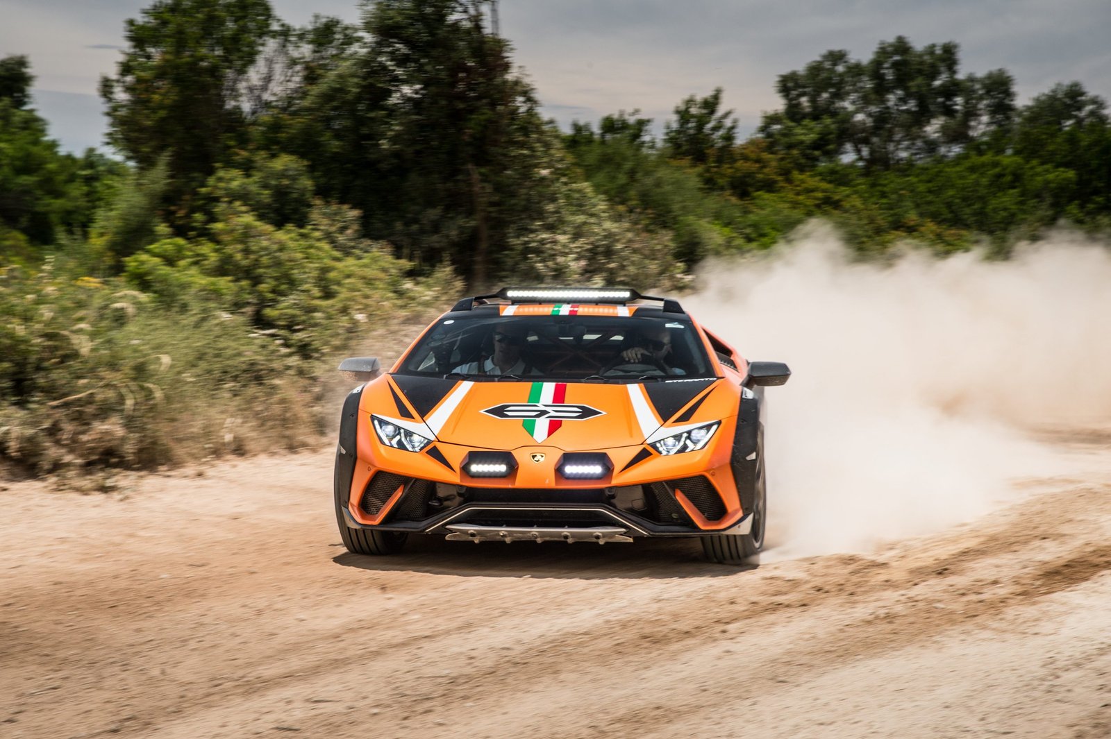 Orange Lamborghini Huracán Sterrato Concept driving head-on down a dirt road, kicking up a massive dust cloud with auxiliary LED lights and front skid plate visible