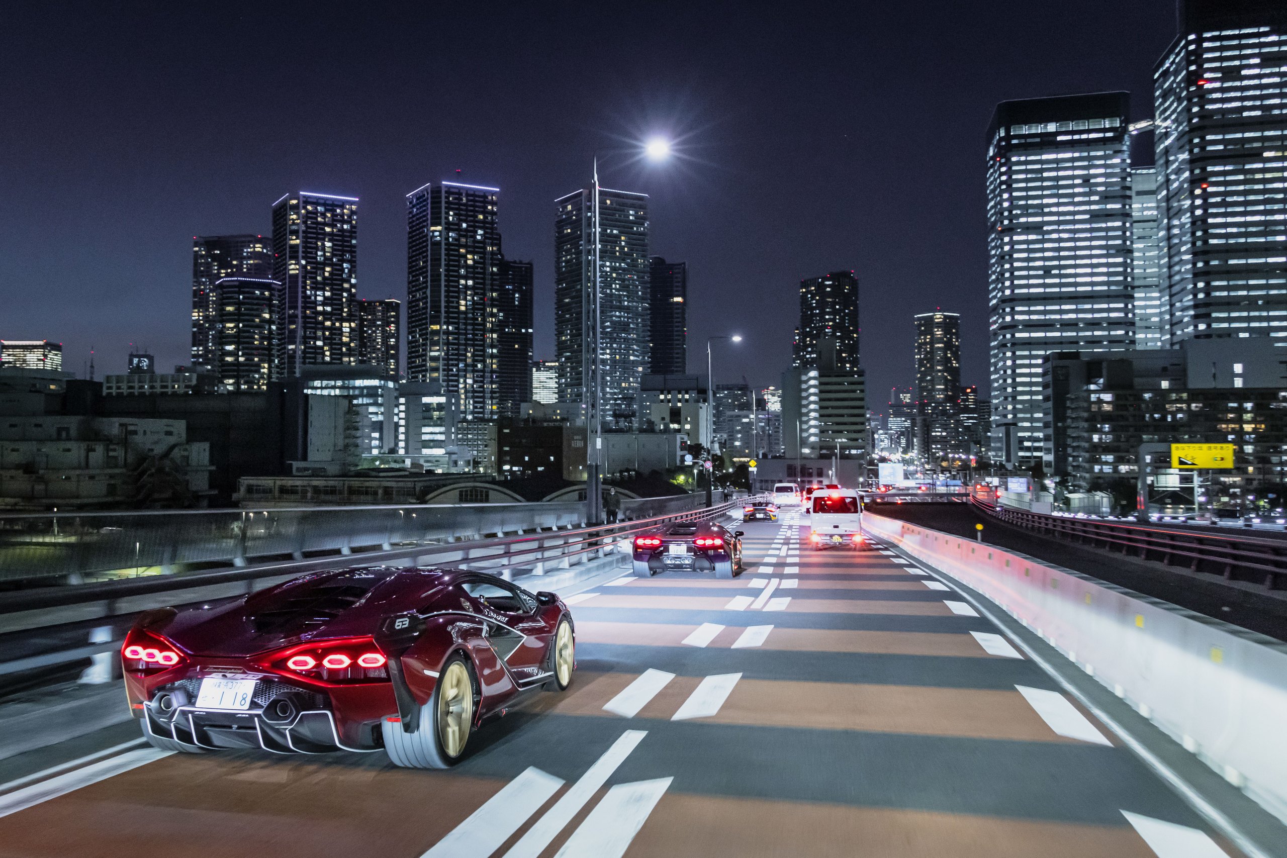 Red lamborghini sián fkp 37 driving on a highway at night with illuminated city skyscrapers in the background
