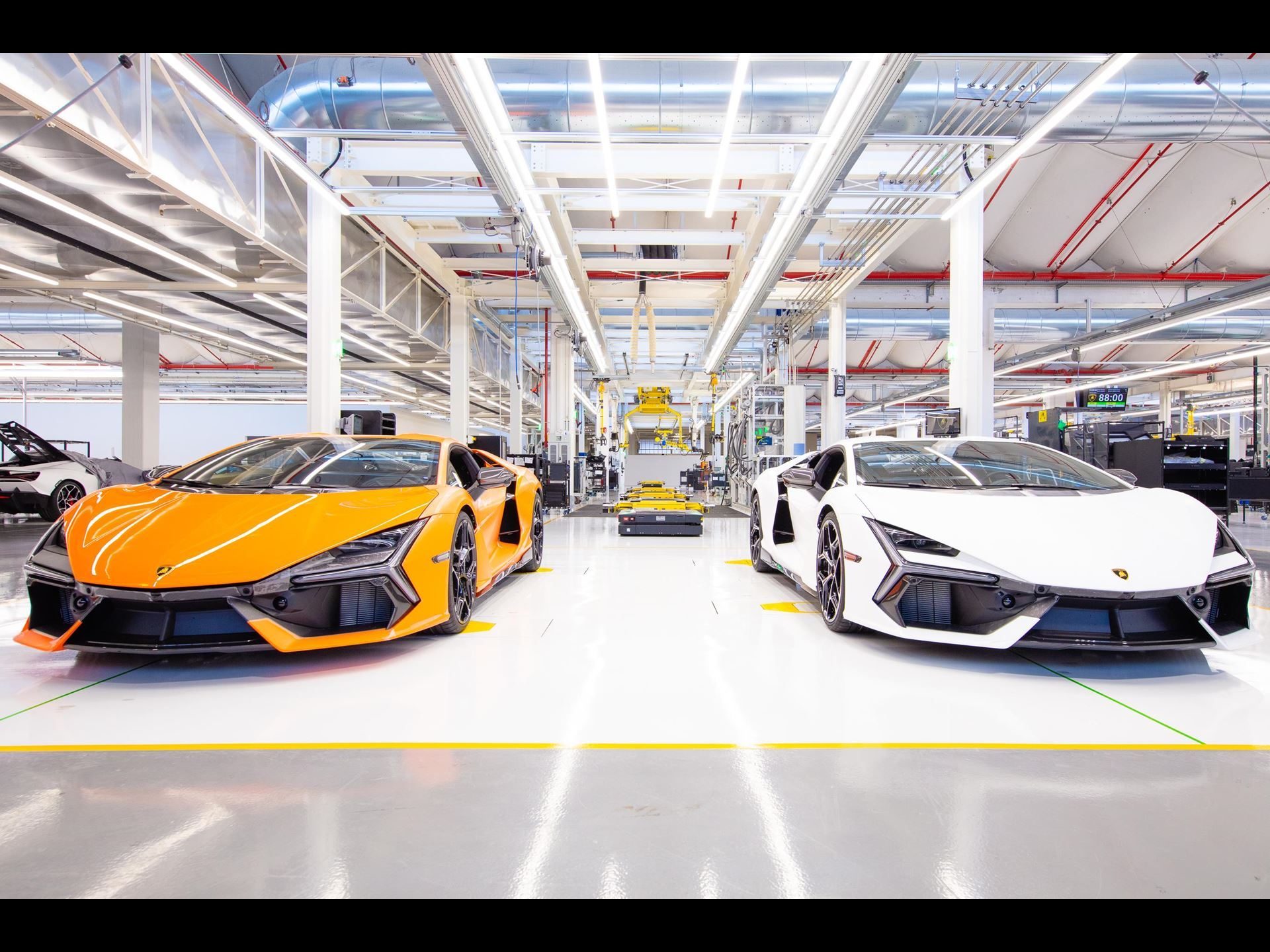 Two lamborghini revuelto supercars, one orange and one white, parked side by side in the sant'agata bolognese manufacturing facility