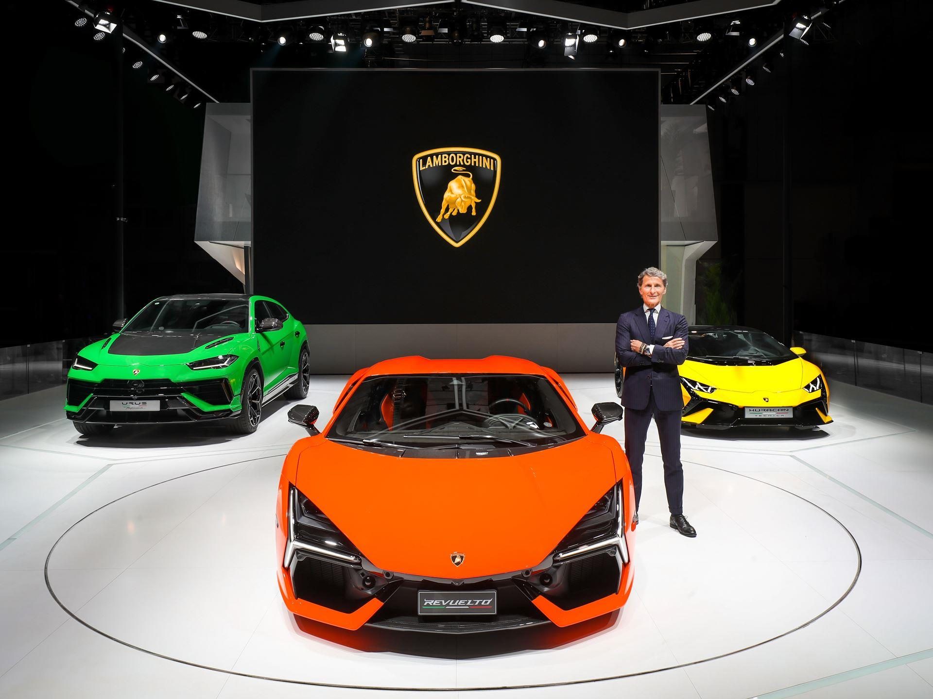 Stephan winkelmann standing in front of an orange revuelto flanked by a green urus and yellow huracán on a stage with lamborghini logo