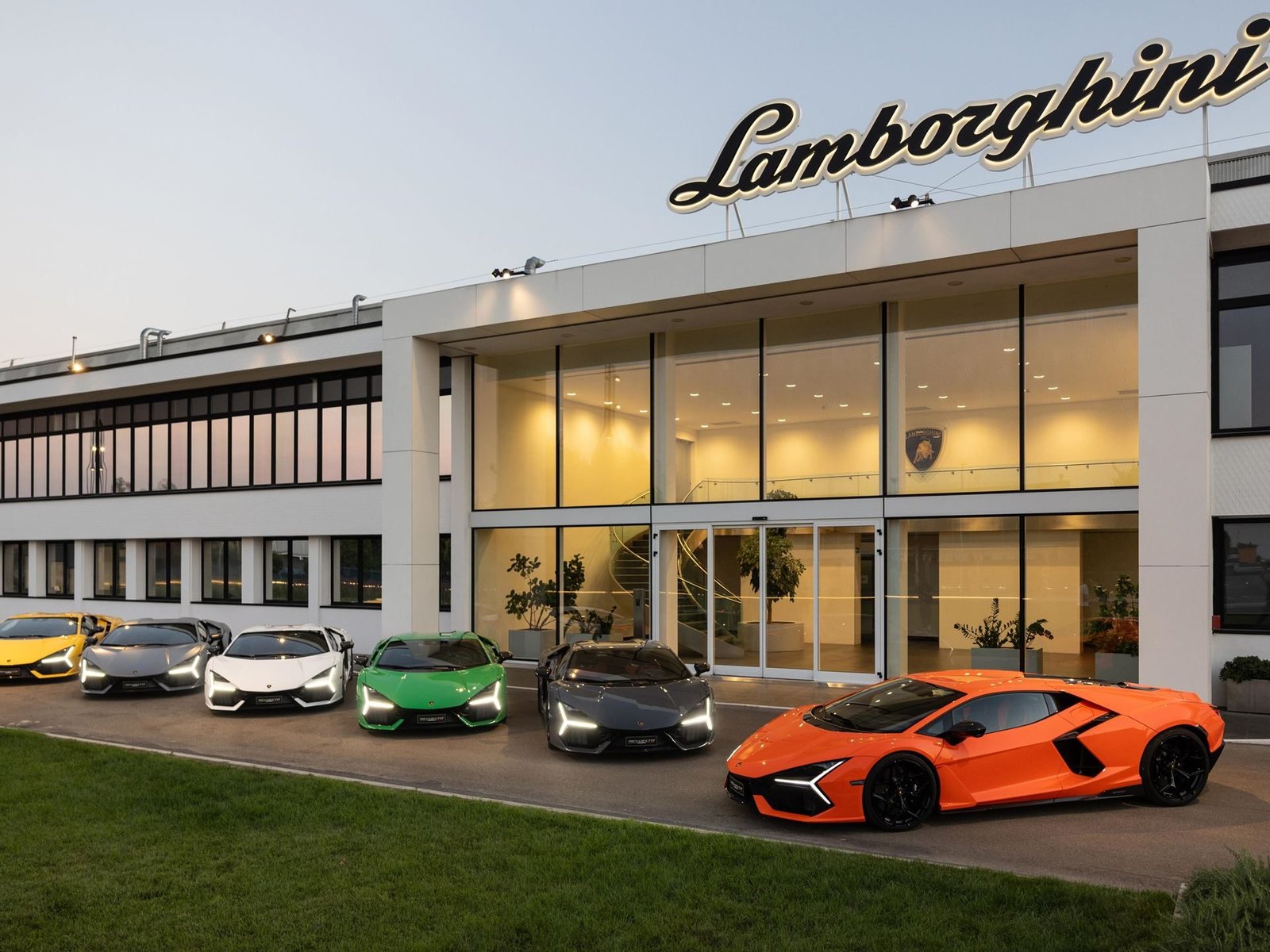 Lineup of six Lamborghini vehicles including an orange Revuelto parked in front of the illuminated Lamborghini factory entrance