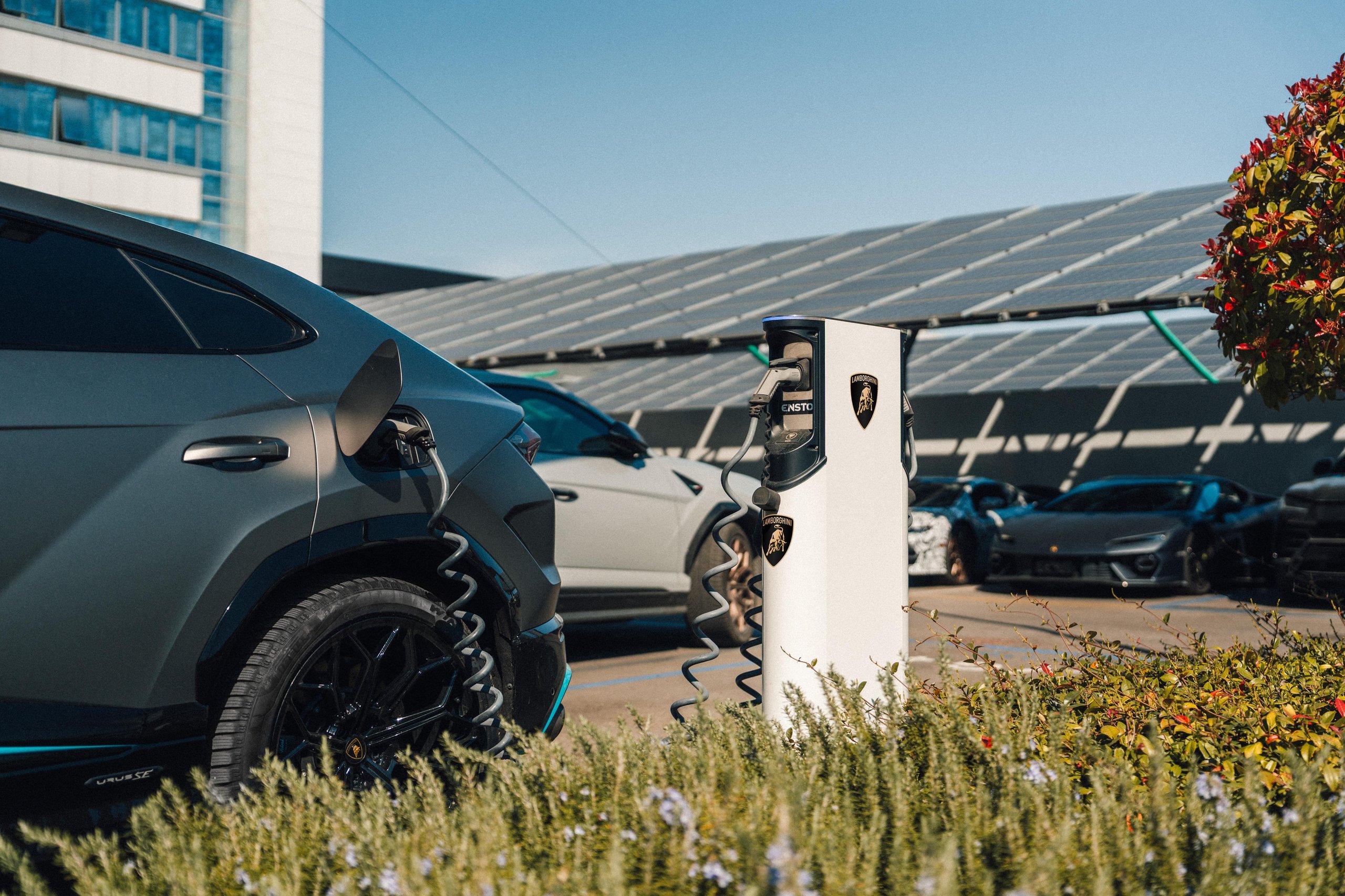 A grey lamborghini urus se charging at a station with solar panels visible in the background and other lamborghini vehicles parked nearby