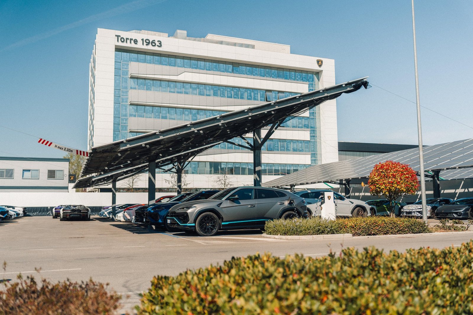 A grey Lamborghini Urus SE charging under a solar panel canopy at the Sant'Agata Bolognese facility, with the Torre 1963 building in the background