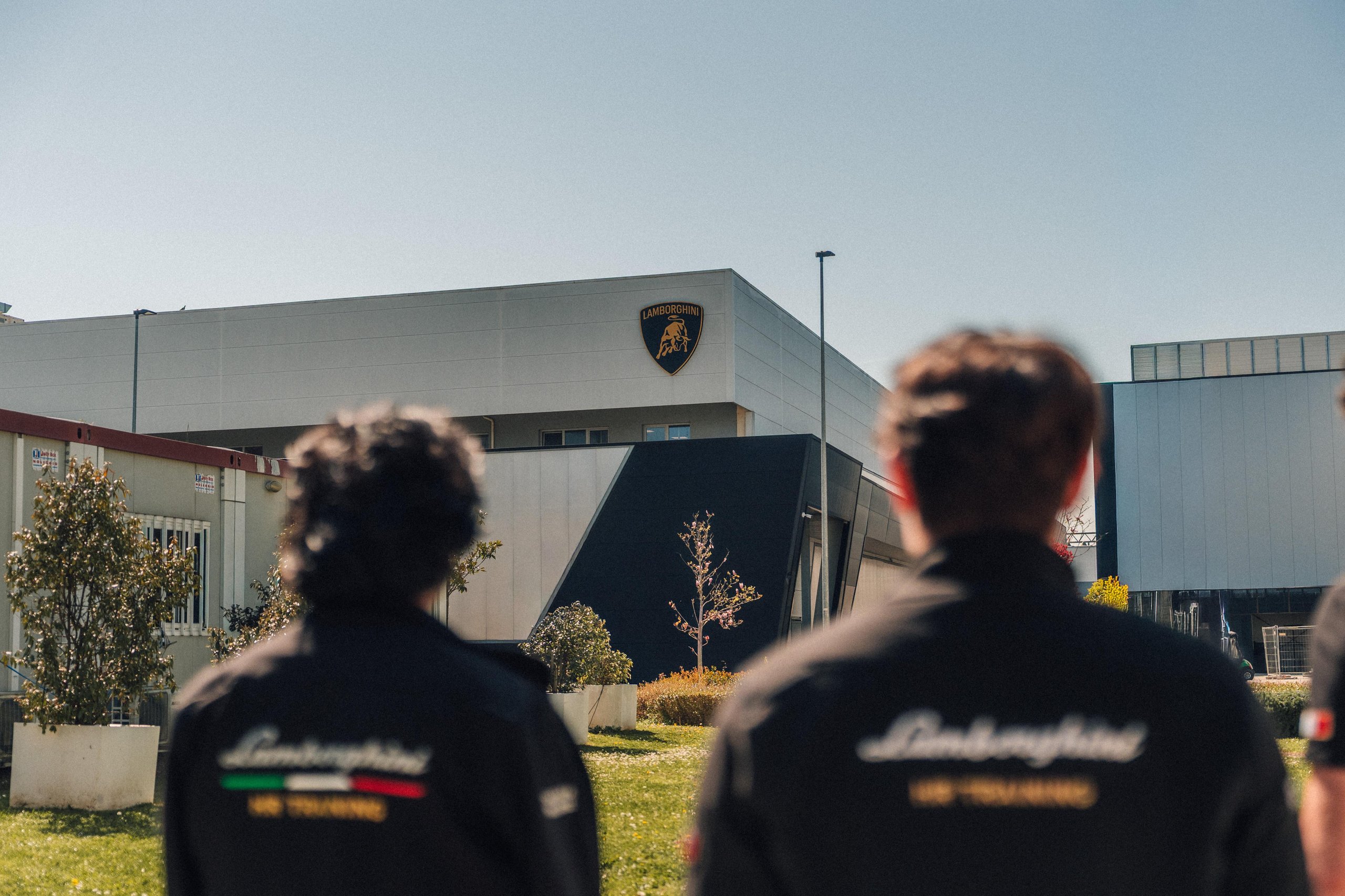 Two individuals in lamborghini young jackets looking toward a modern lamborghini building under a clear blue sky