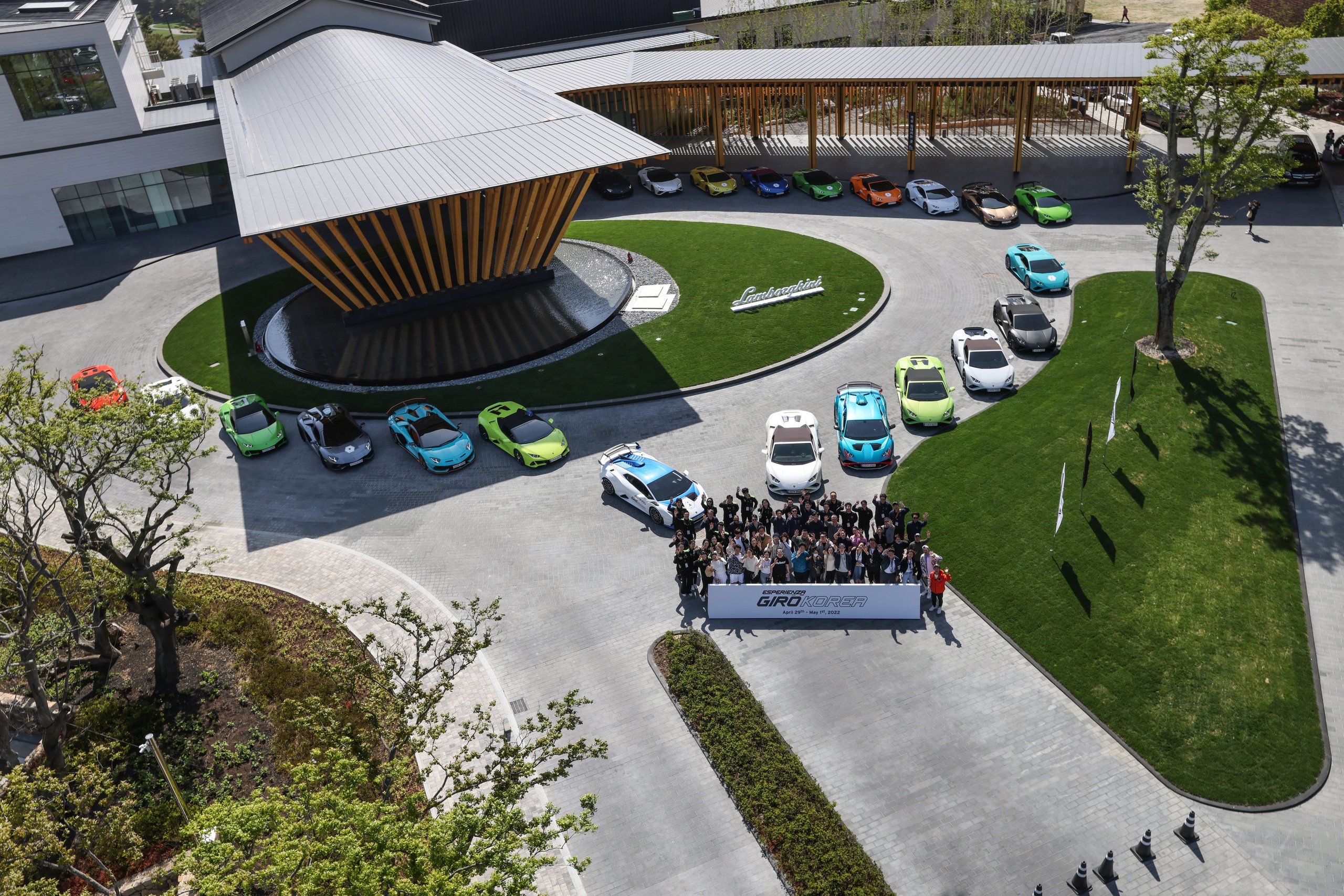 Aerial view of a large gathering of diverse lamborghini vehicles and owners holding a banner in front of a modern building