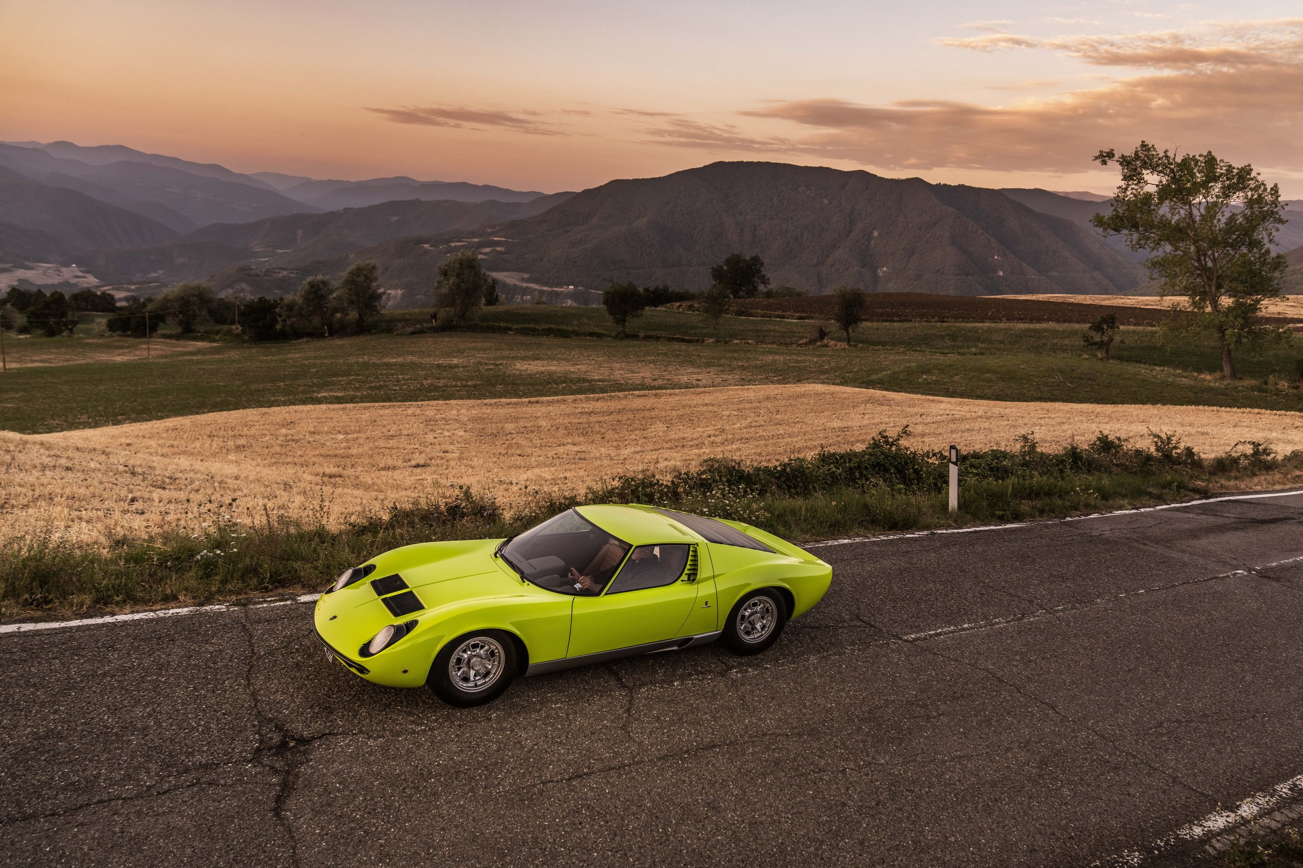 A vibrant lime green lamborghini miura driving on a winding country road at sunset with rolling hills in the background