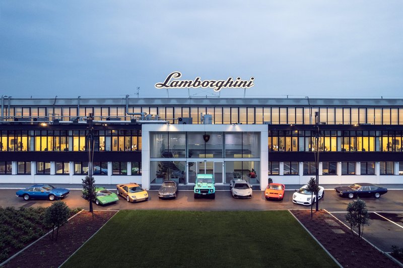 A diverse lineup of classic and modern Lamborghini vehicles arranged in front of the Lamborghini factory at twilight, spanning decades of the brand's history