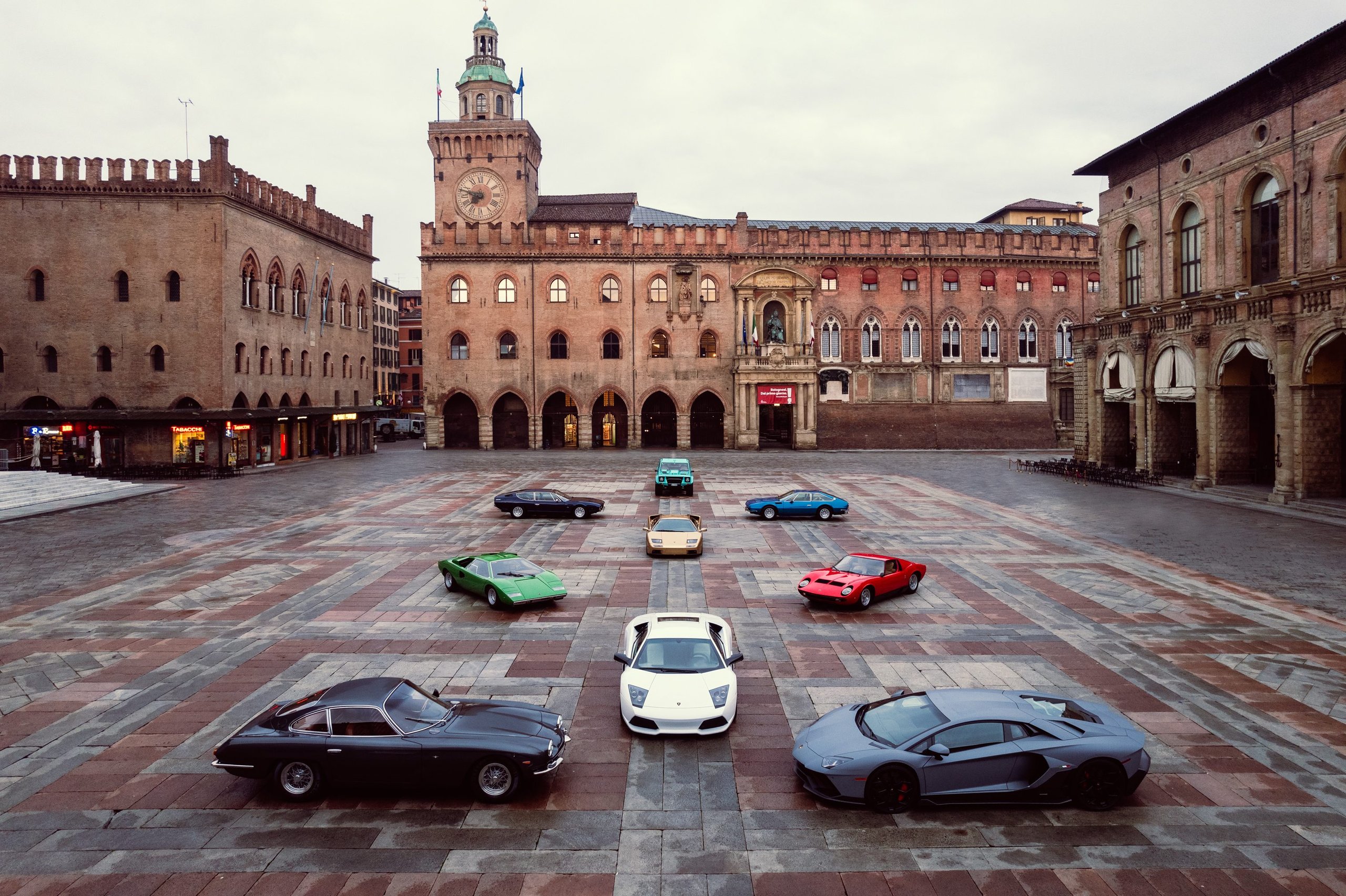 A diverse collection of lamborghini vehicles spanning different eras arranged in a historic italian city square with grand architecture