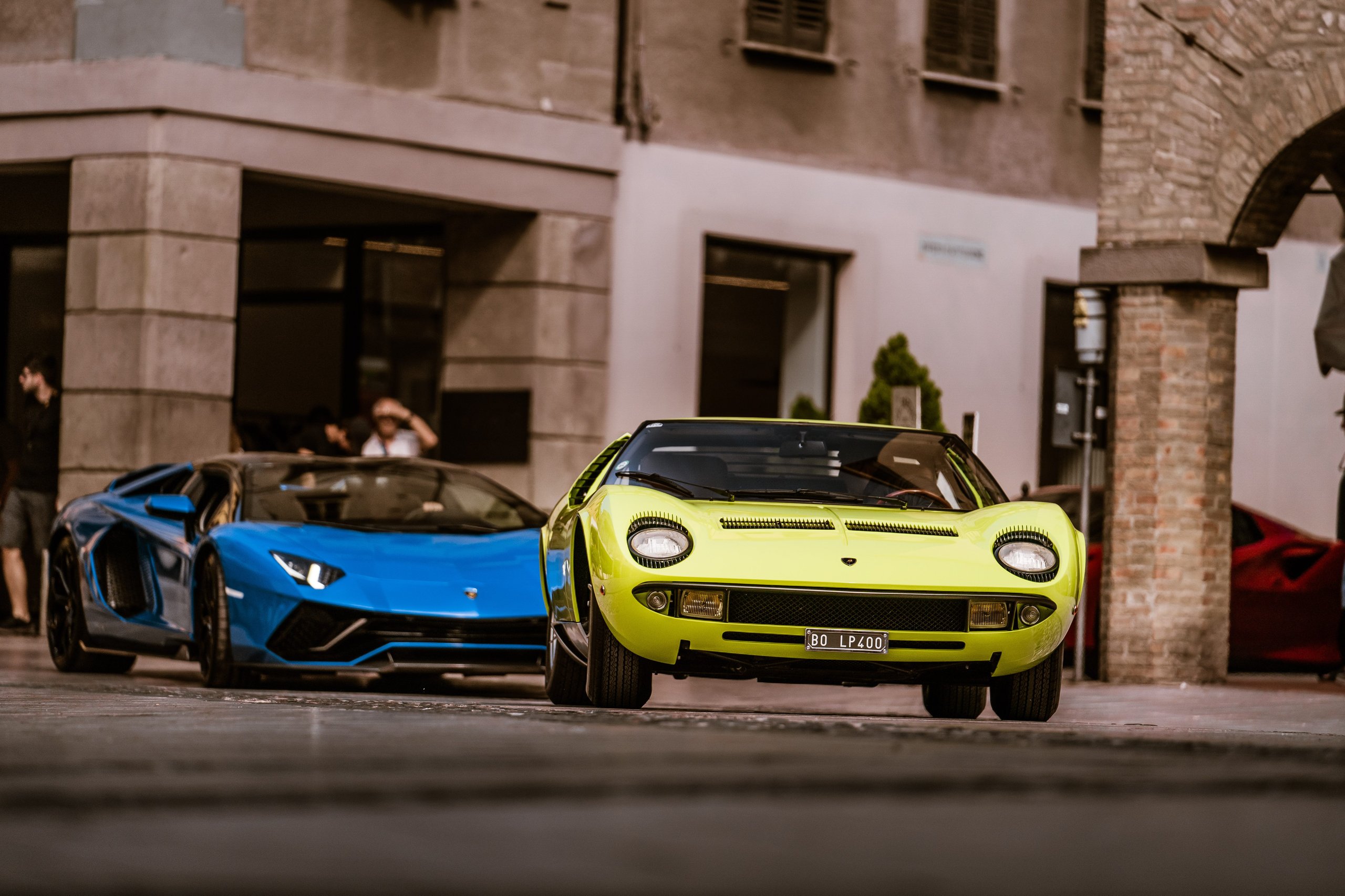 A classic yellow lamborghini miura leads a modern blue lamborghini aventador down a narrow european street