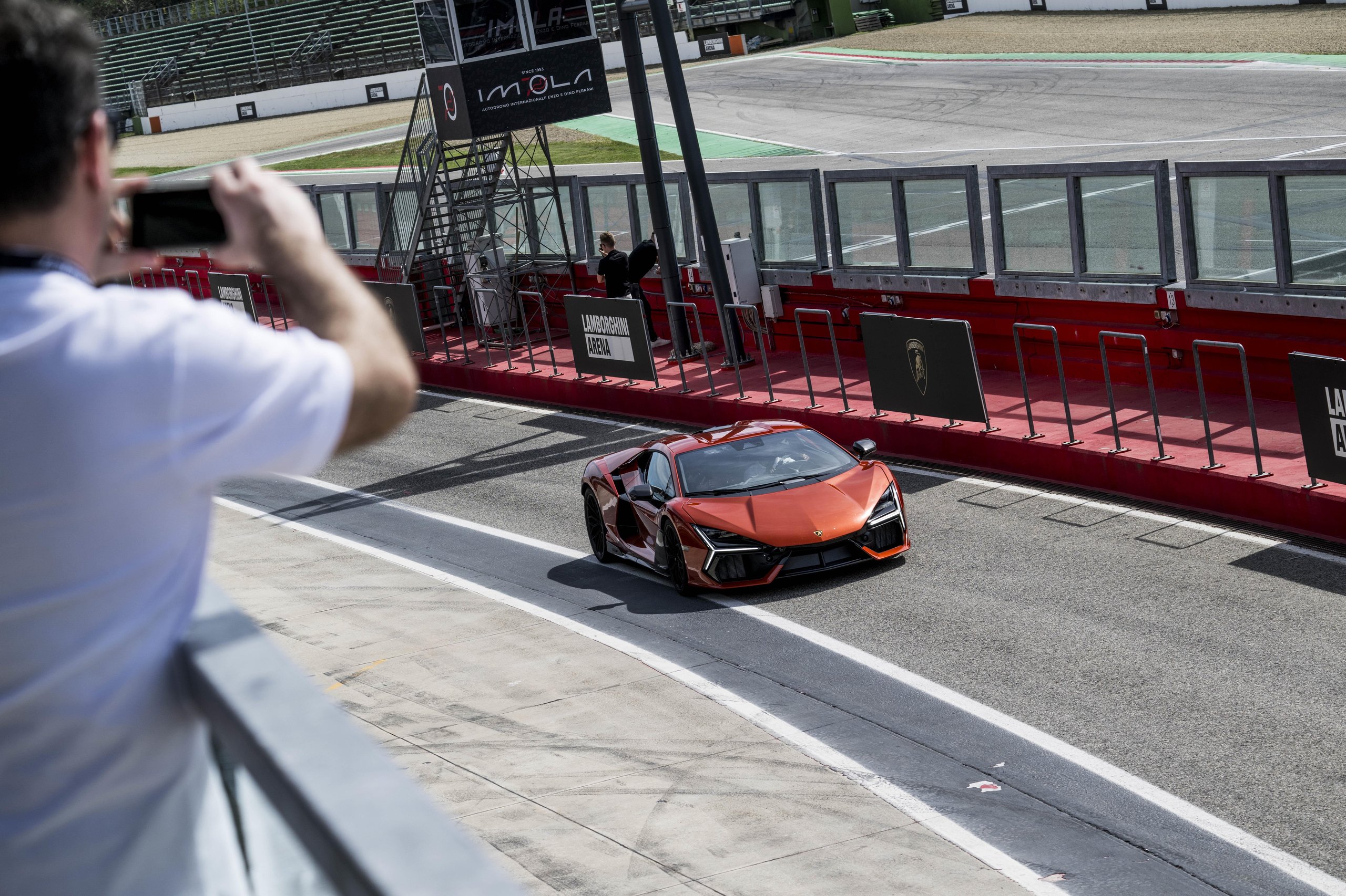 Lamborghini Arena 2026 Returns To Imola: Why The Brand'S Biggest Owner Gathering Matters More This Time 9 An orange lamborghini revuelto driving on the imola racetrack viewed from an elevated perspective with a fan photographing in the foreground