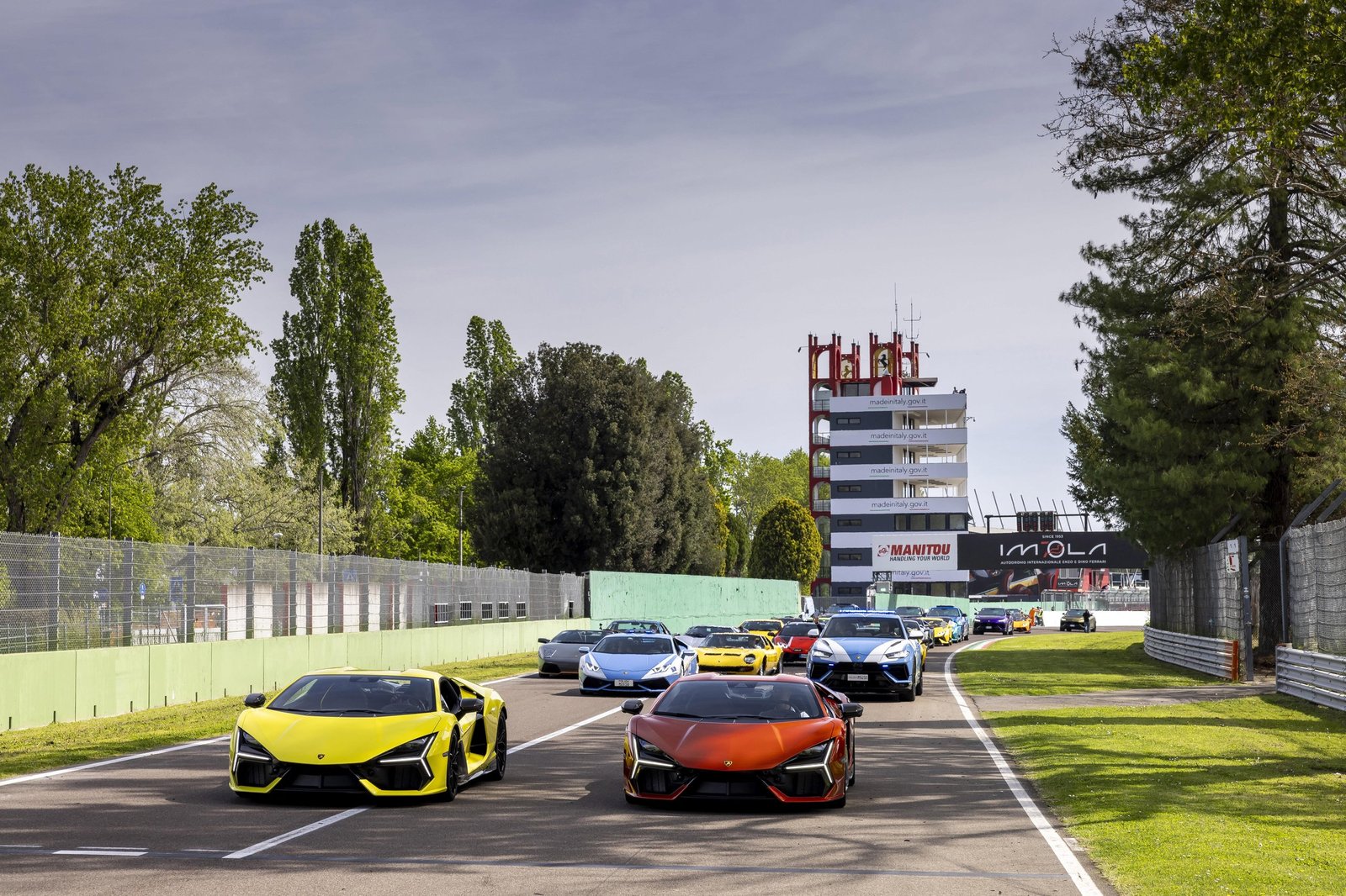 A procession of Lamborghini Revuelto, Huracán, and Urus models driving on the Imola Circuit during the Lamborghini Arena event