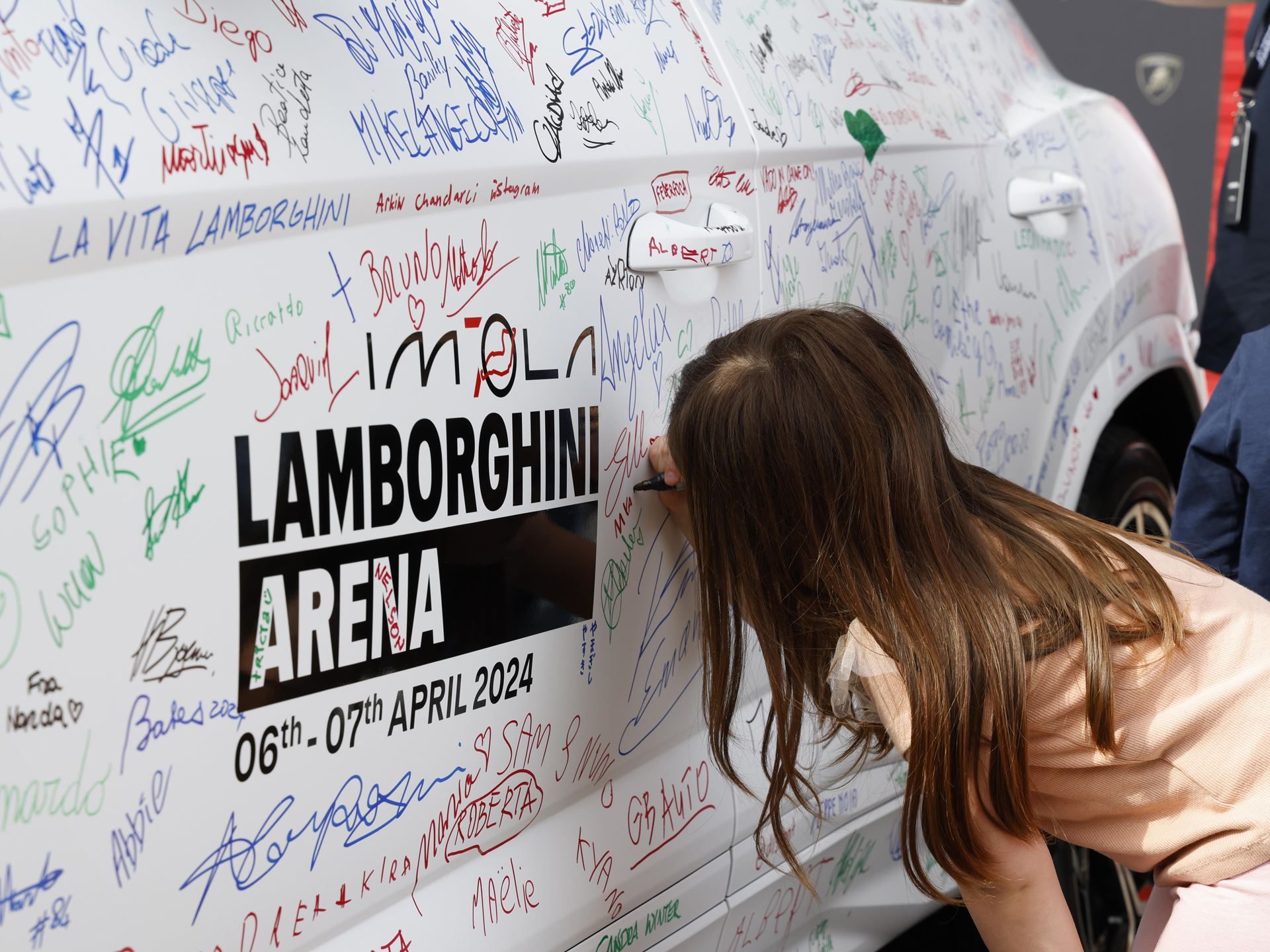 Lamborghini Arena 2026 Returns To Imola With Super Trofeo Racing, Miura Anniversary, And A Bigger Playbook 11 A young girl signs the side of a white lamborghini urus covered in signatures and lamborghini arena 2024 branding