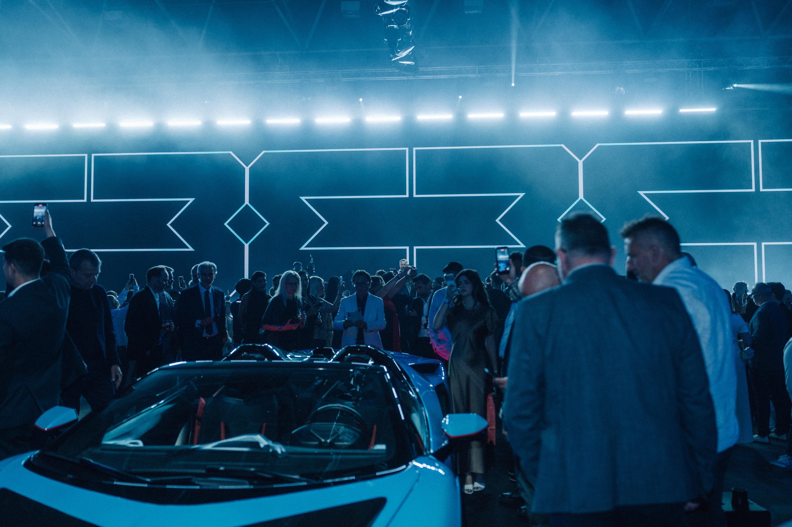 Why Lamborghini Hired Benny Benassi To Turn Arena Into A Homecoming For 6,000 Owners 3 A light blue lamborghini revuelto surrounded by a crowd of attendees at lamborghini arena, with blue geometric lighting in the background