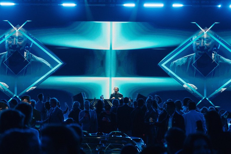 DJ Benny Benassi performing on stage at Lamborghini Arena with large screens and blue geometric lighting, seen from behind a crowd of attendees