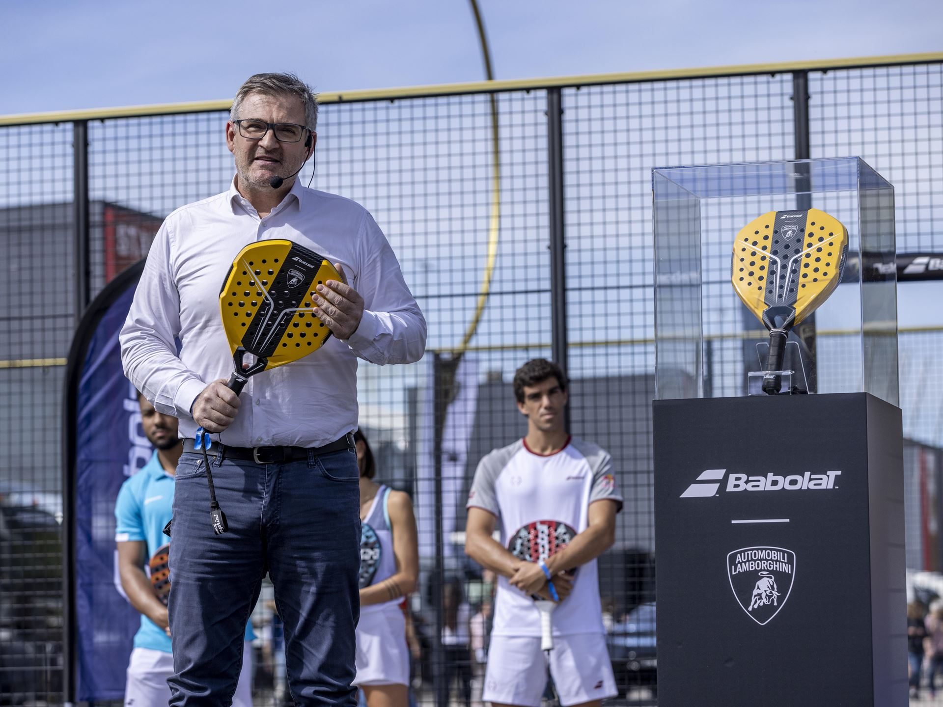 Lamborghini Builds 50 Padel Racquets In Its Supercar Factory, And That'S The Point 7 Speaker holding the yellow and black bl001 padel racquet at the lamborghini arena launch event, with a display case visible behind