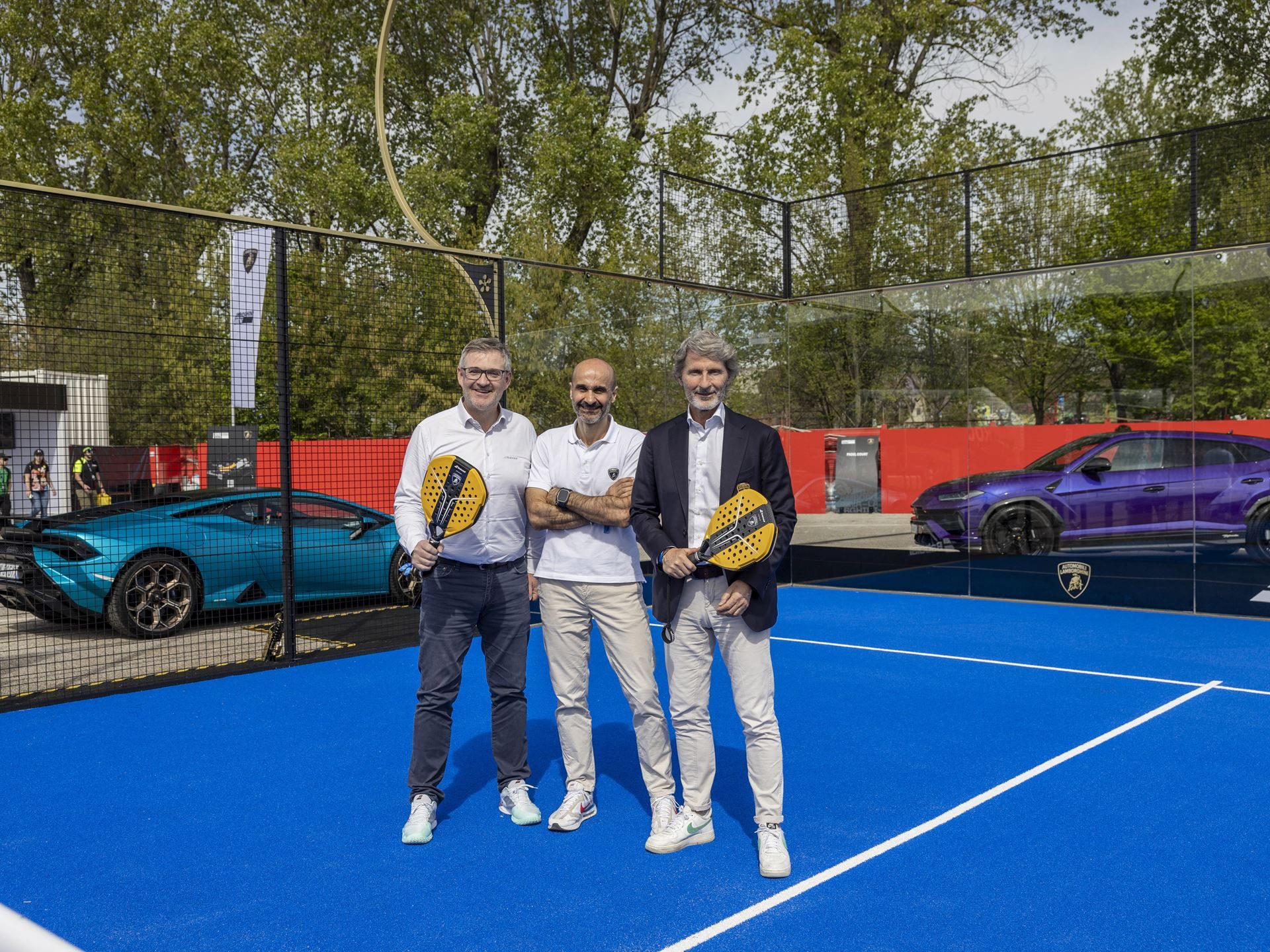 Lamborghini Builds 50 Padel Racquets In Its Supercar Factory, And That'S The Point 9 Three men holding babolat x lamborghini padel racquets on a blue court with a teal huracán and purple urus parked behind them