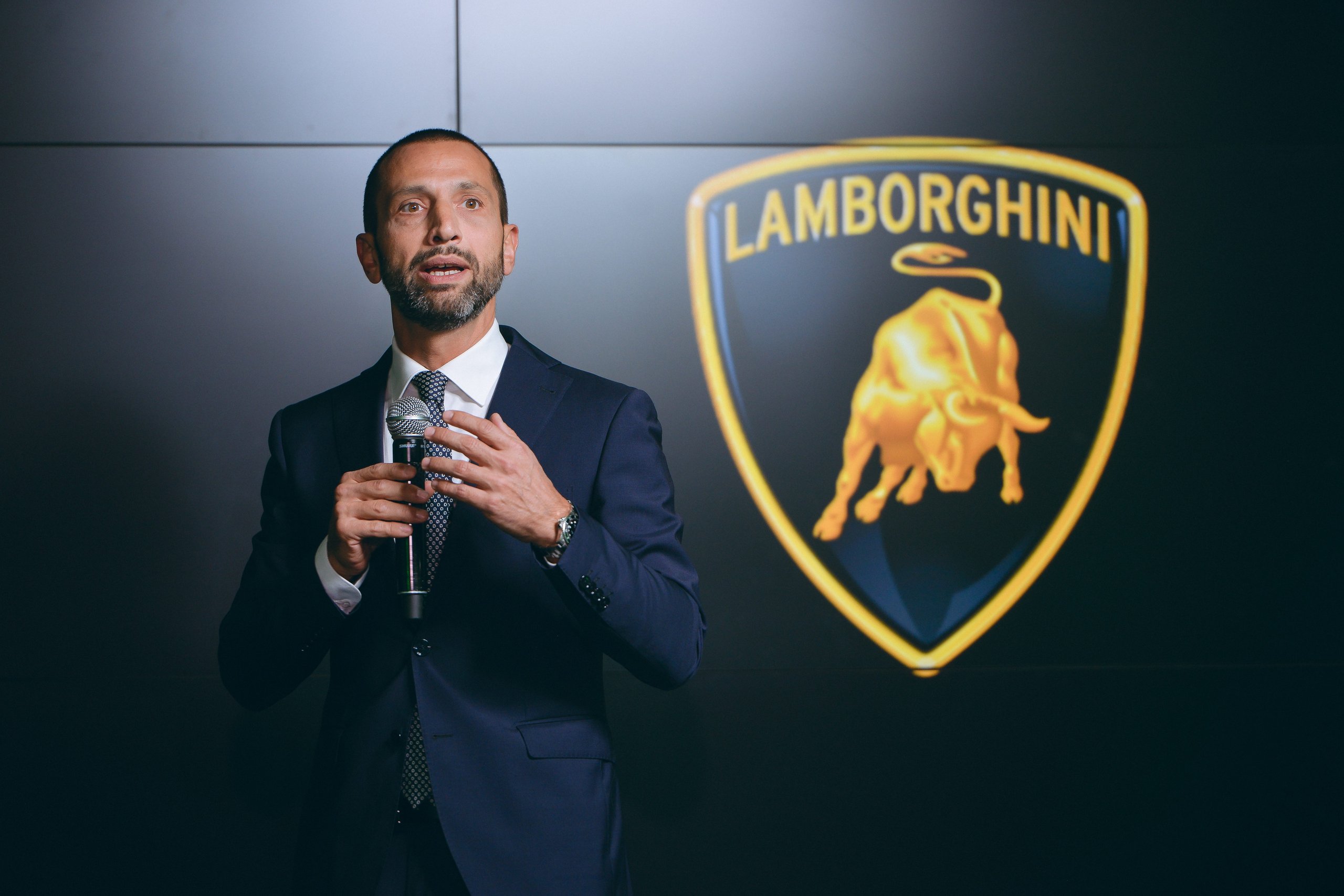 Lamborghini Baku: Why A 225 Square Meter Showroom Signals Serious Ambitions For The Transcaucasia Region 9 A speaker in a suit addresses the audience at the lamborghini baku opening with an illuminated lamborghini shield logo behind him