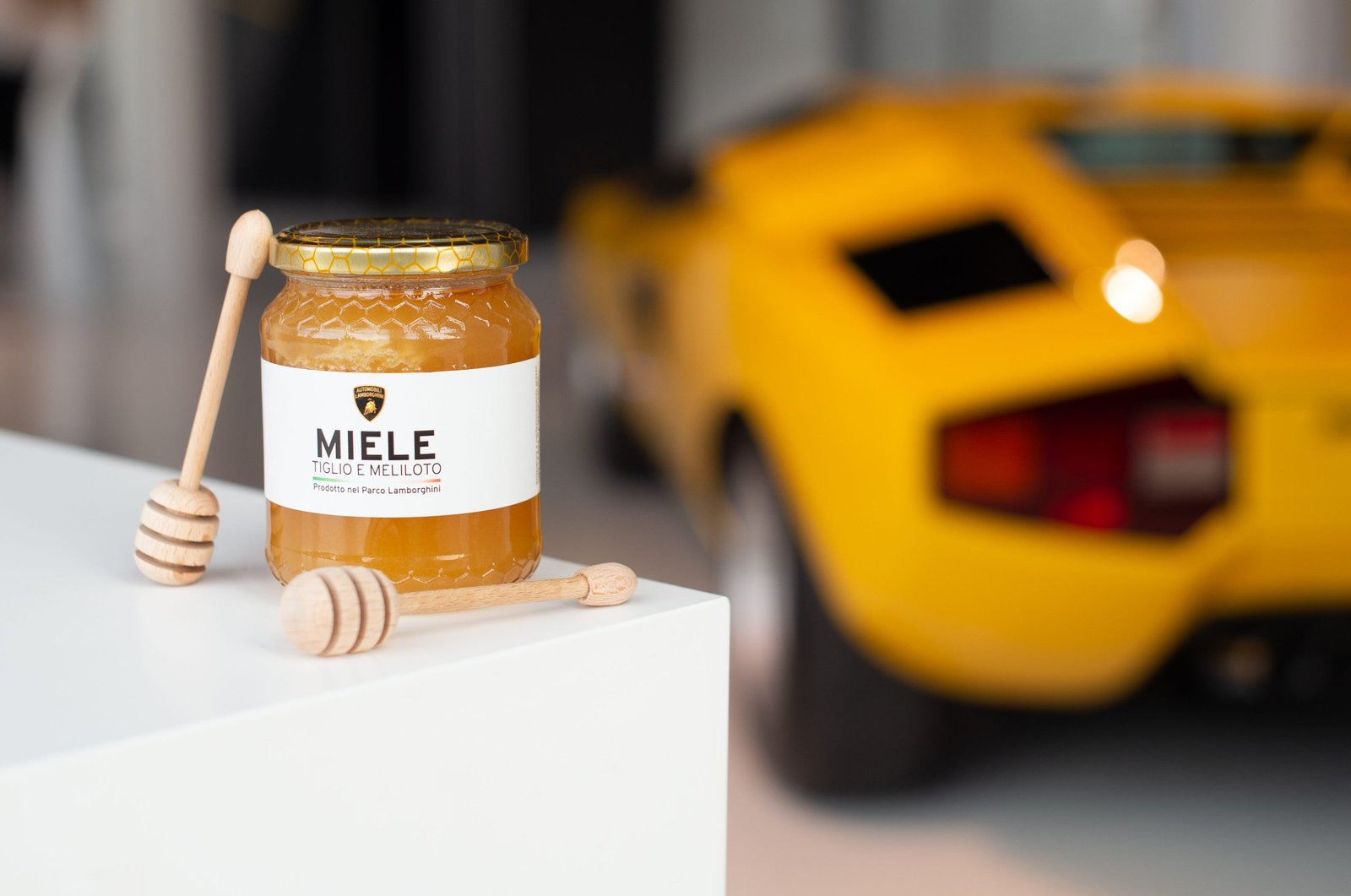 A jar of Lamborghini-branded honey with wooden dippers, a yellow Lamborghini Countach blurred in the background