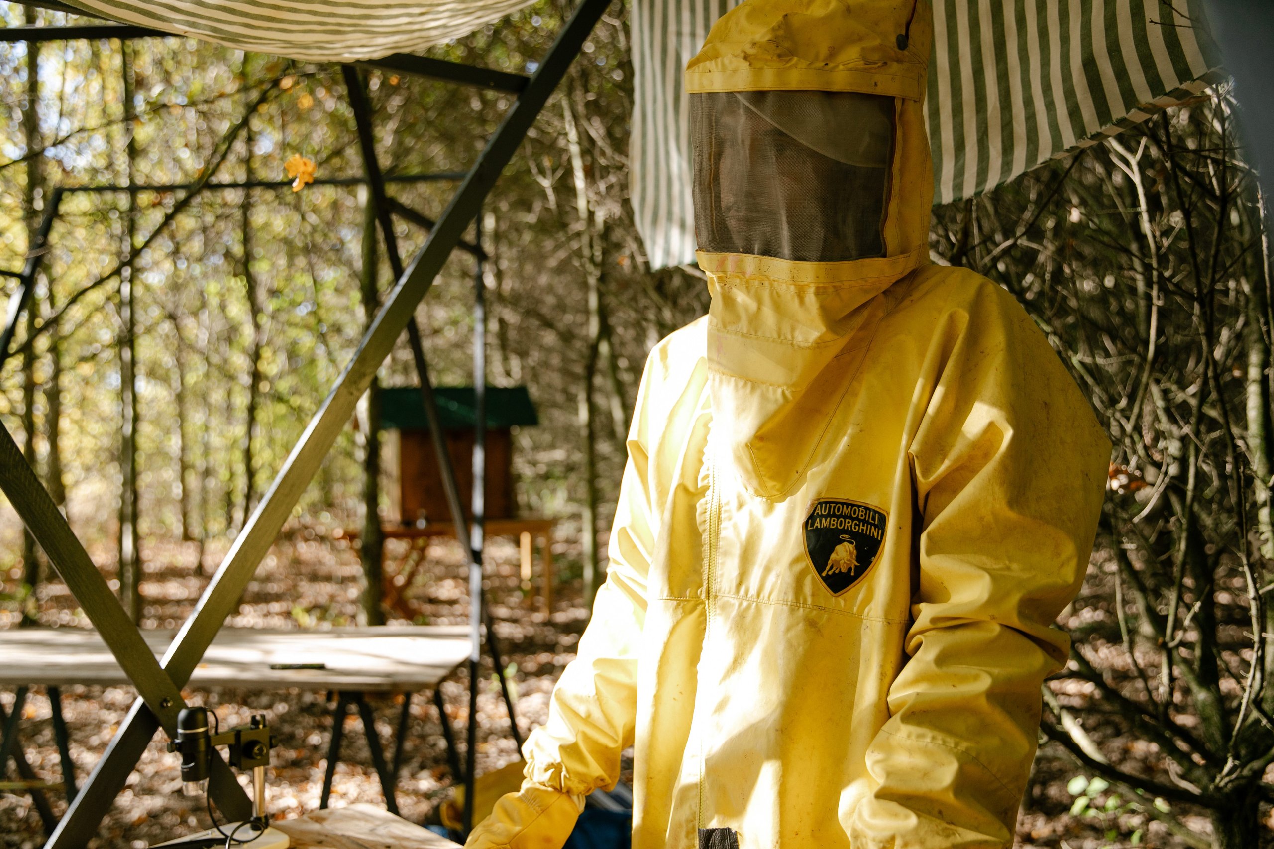 600,000 Bees And Sim Cards: How Lamborghini Turned Its Factory Grounds Into A Living Environmental Lab 3 A person wearing a yellow beekeeper suit bearing an automobili lamborghini patch on the chest, standing in a wooded outdoor setting