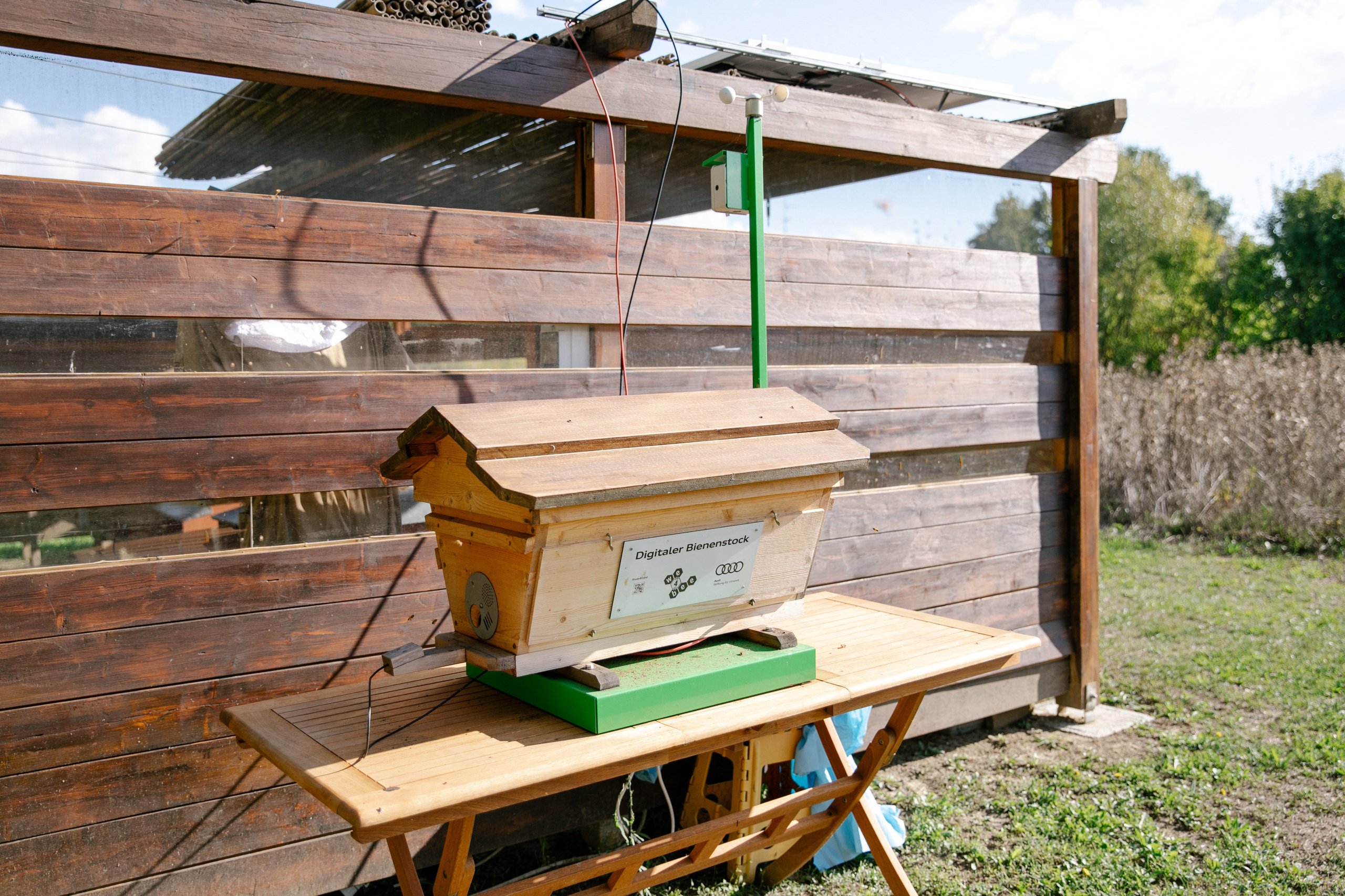 600,000 Bees And Sim Cards: How Lamborghini Turned Its Factory Grounds Into A Living Environmental Lab 5 A wooden beehive with a green base displaying digitaler bienenstock signage and audi logos, placed on a wooden table outdoors