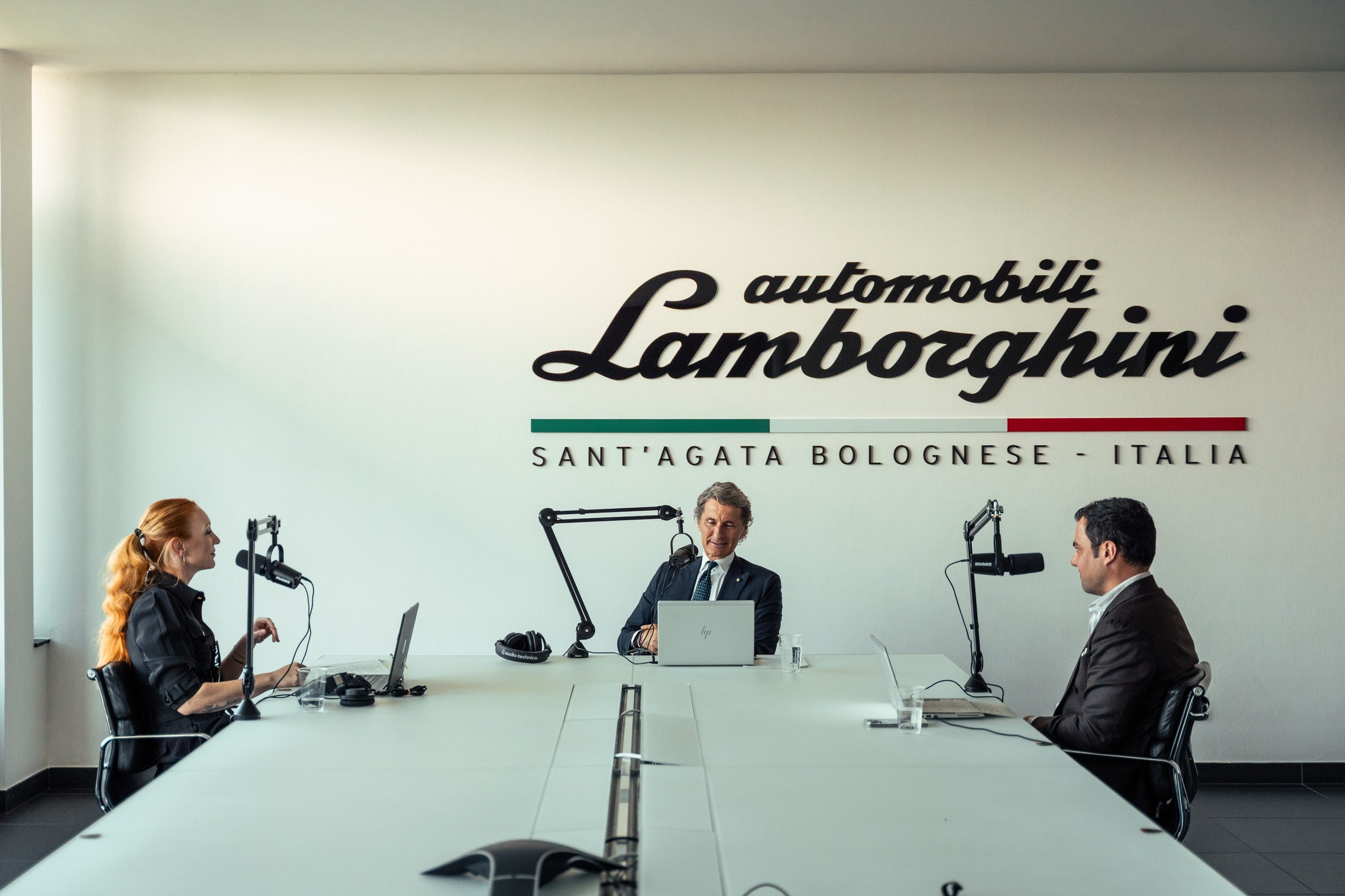 Three individuals seated at a white table in a recording studio with lamborghini branding on the wall behind them