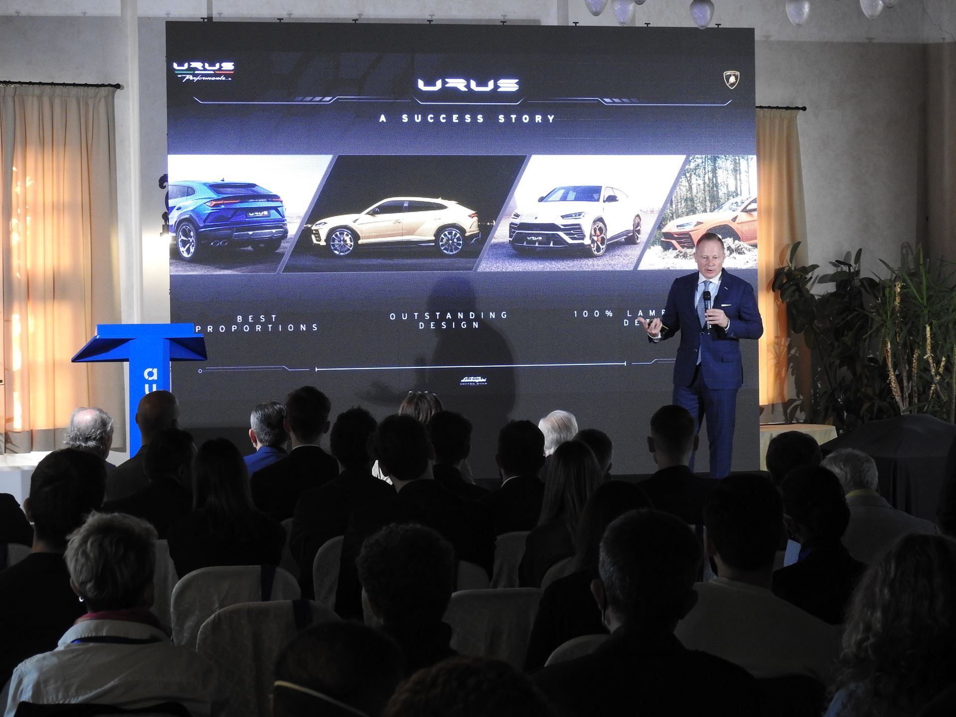 Mitja Borkert Used The Autostyle Stage To Explain Why Every Lamborghini Starts With A Single Line 7 Speaker presenting on stage with large screen showing urus a success story and images of lamborghini urus models