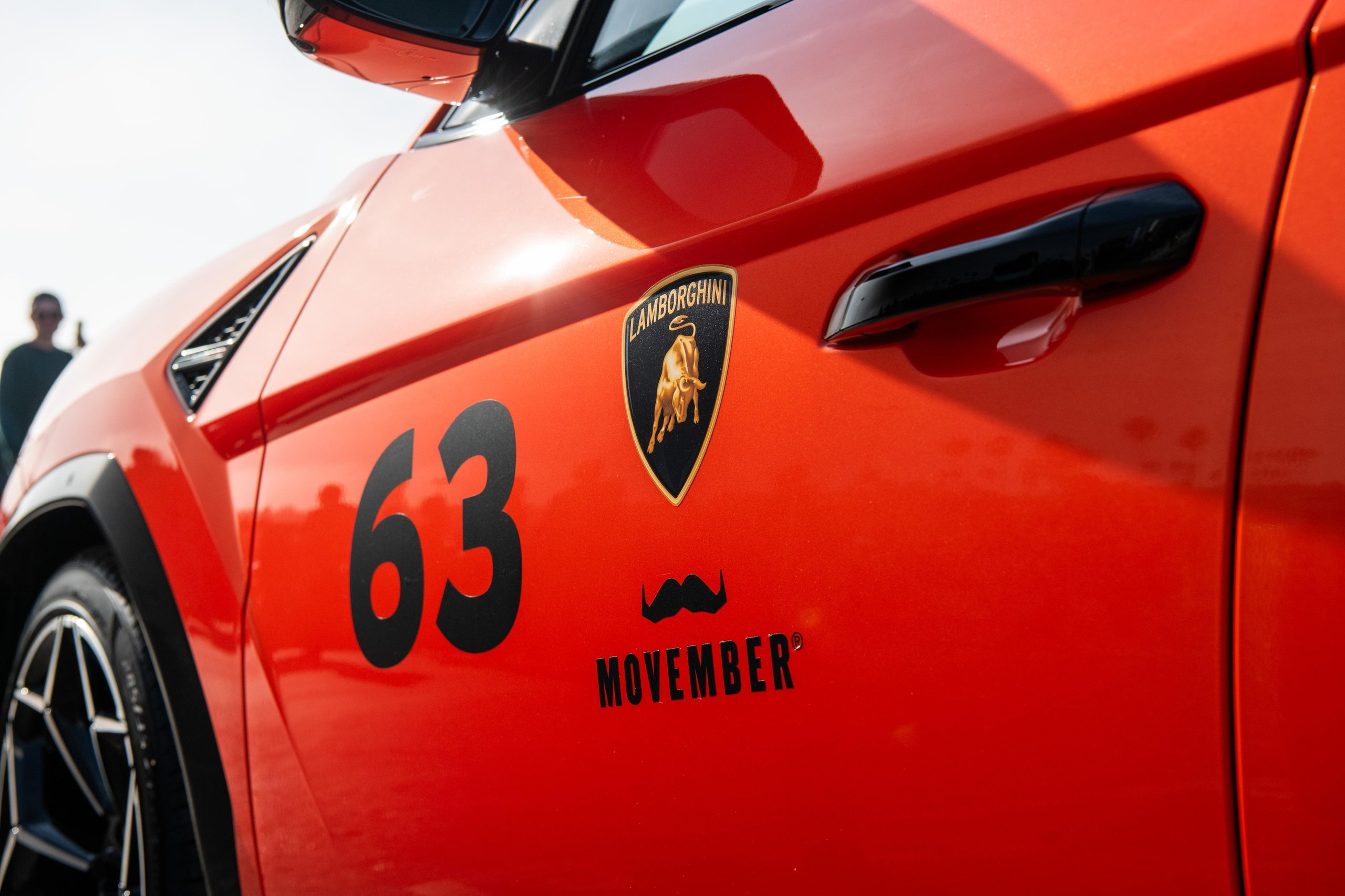 200 Mustache-Wearing Lamborghinis Took Over Malibu, And The Ceo Was Leading The Pack 5 Close-up detail of an orange lamborghini urus showing the lamborghini shield emblem, number 63, and movember mustache logo