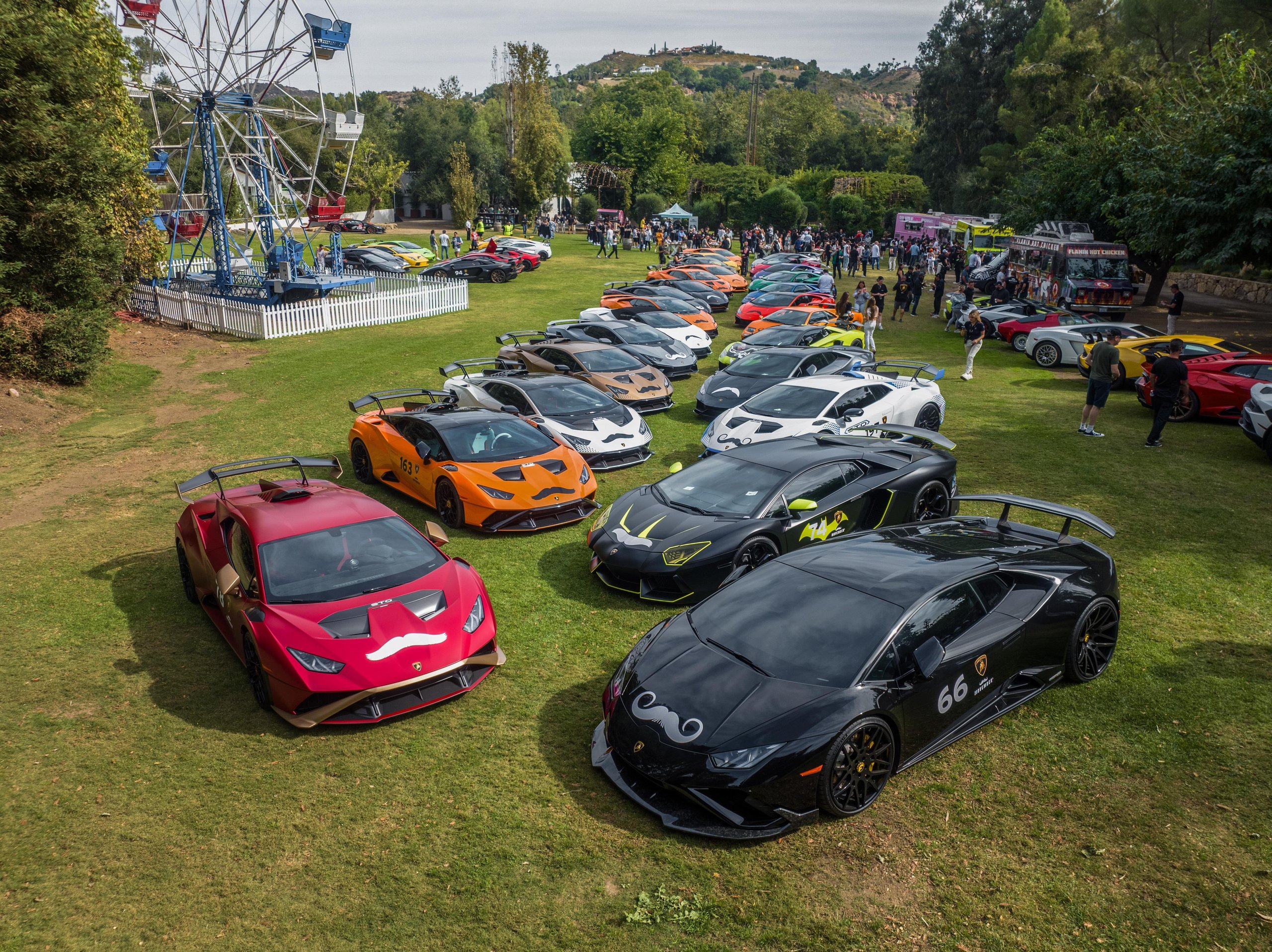 200 Mustache-Wearing Lamborghinis Took Over Malibu, And The Ceo Was Leading The Pack 11 Elevated view of a large gathering of lamborghini supercars with movember decals on a green lawn with a ferris wheel and event tents in the background