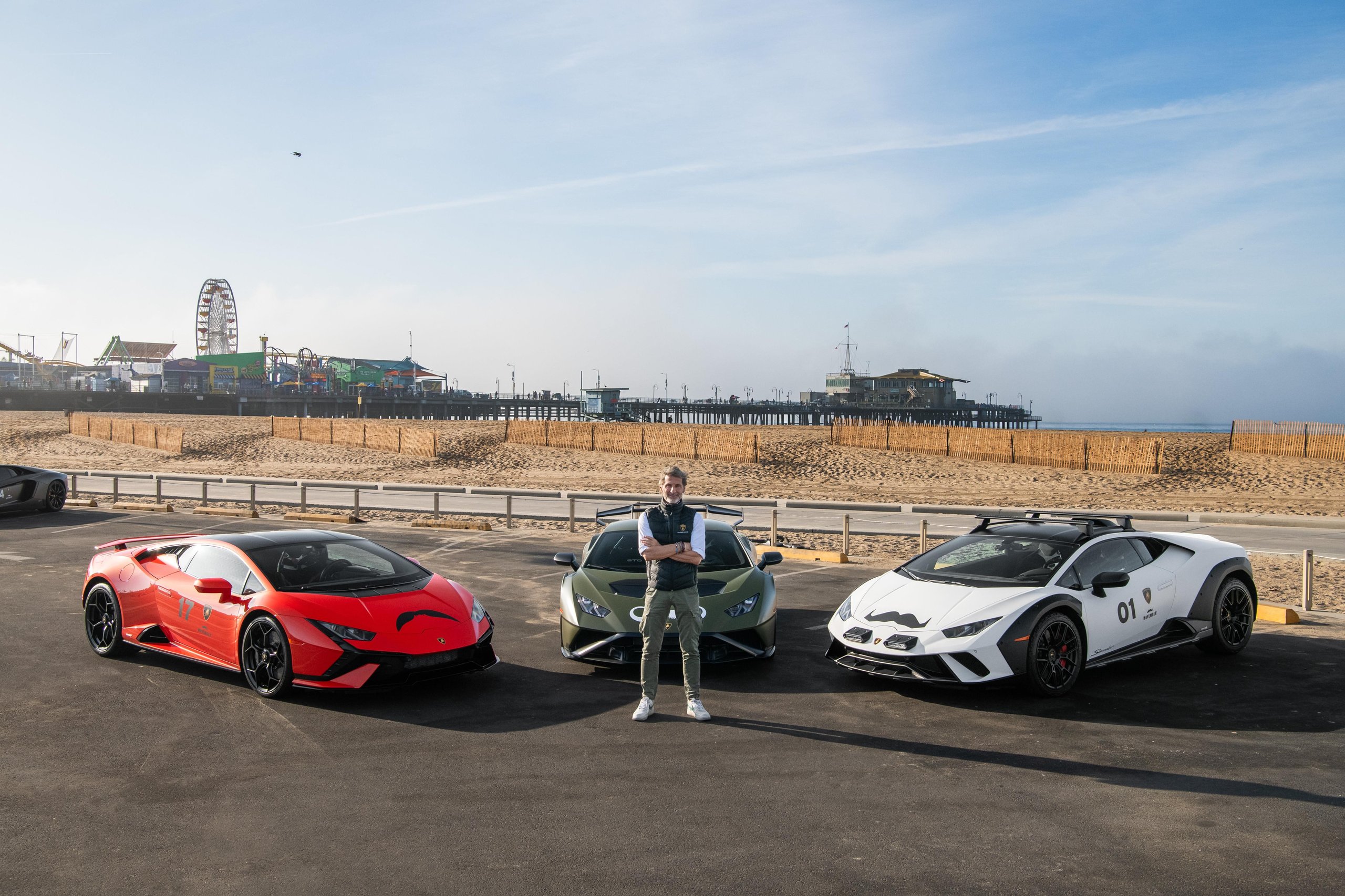 200 Mustache-Wearing Lamborghinis Took Over Malibu, And The Ceo Was Leading The Pack 7 A man stands between a red huracán sto, green huracán sterrato, and white huracán evo near santa monica pier with a ferris wheel in the background